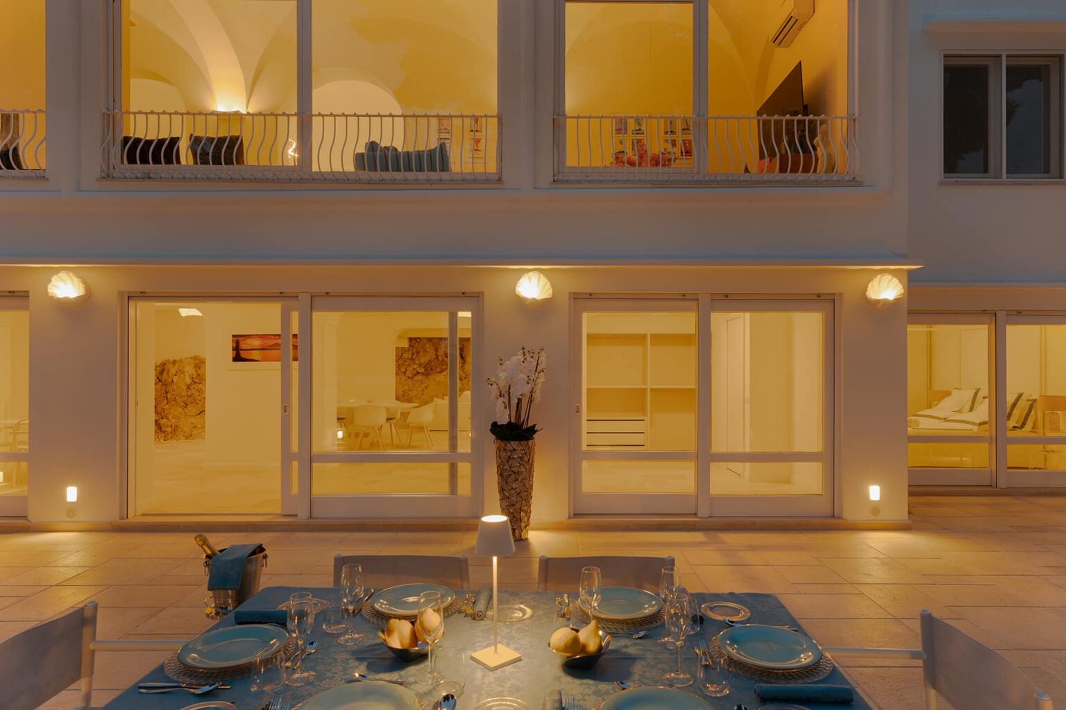 Elegant outdoor dining setup in front of a modern villa with large glass windows, showcasing warm interior lighting and stylish decor. | Only on SopranoVillas