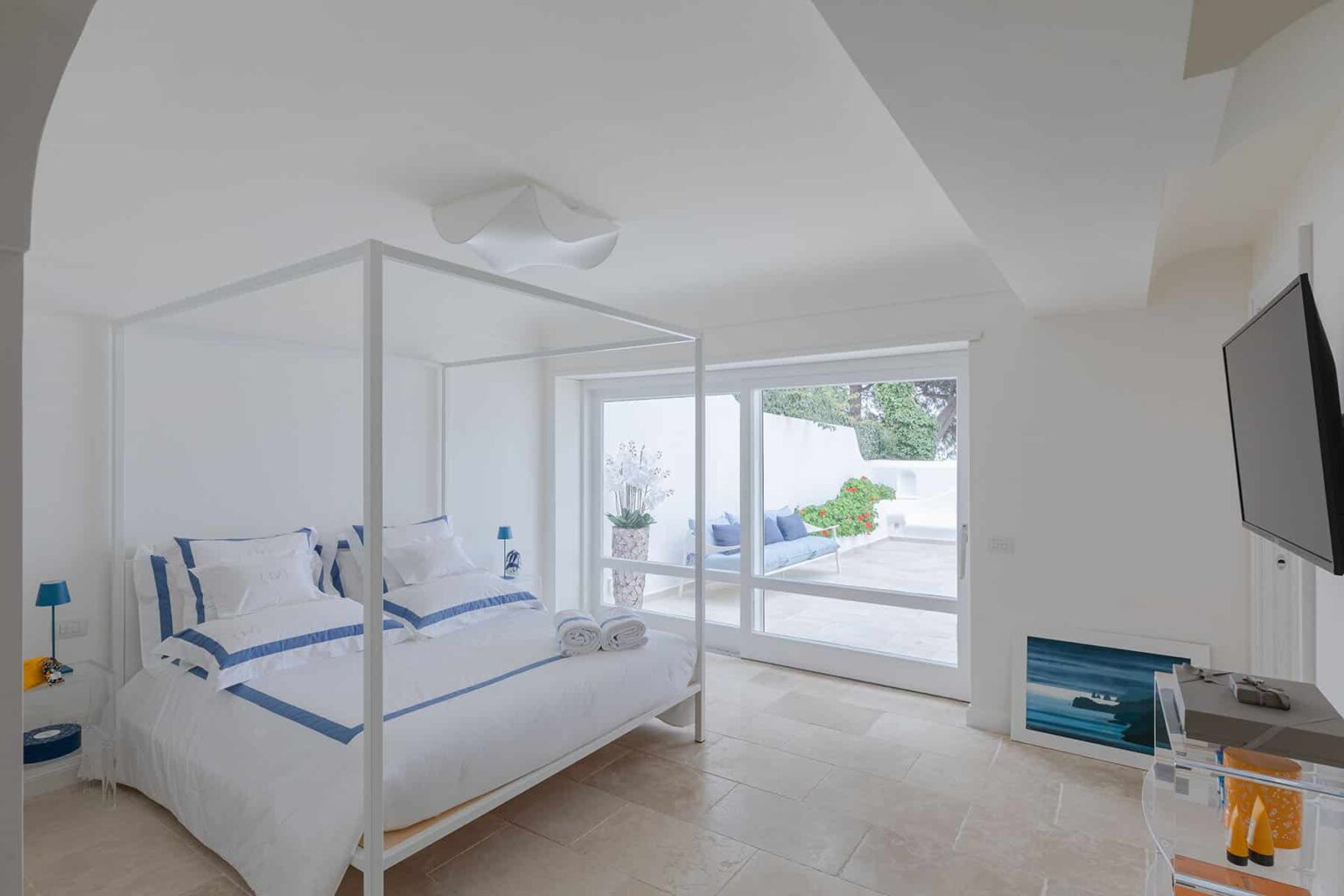 Bright and airy bedroom featuring a four-poster bed with white linens, large windows offering natural light, and minimalist decor. | Only on SopranoVillas