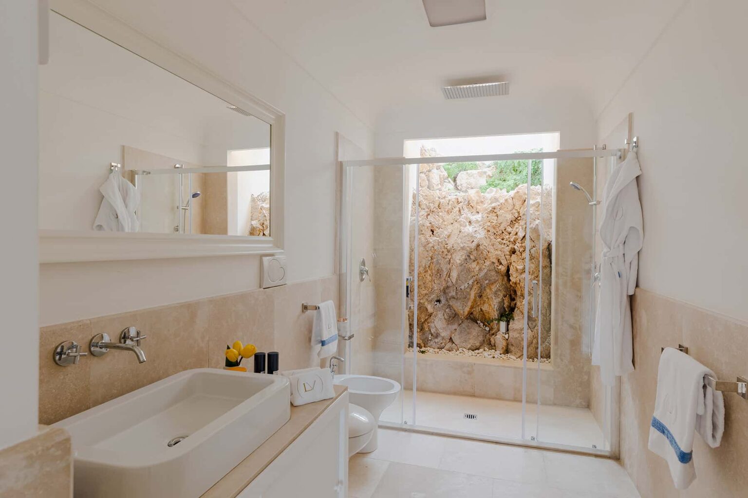 Modern bathroom featuring a spacious shower area with a natural rock wall, a large mirror above a sleek sink, and minimalist decor. Bright natural light illuminates the space through a window. | Only on SopranoVillas