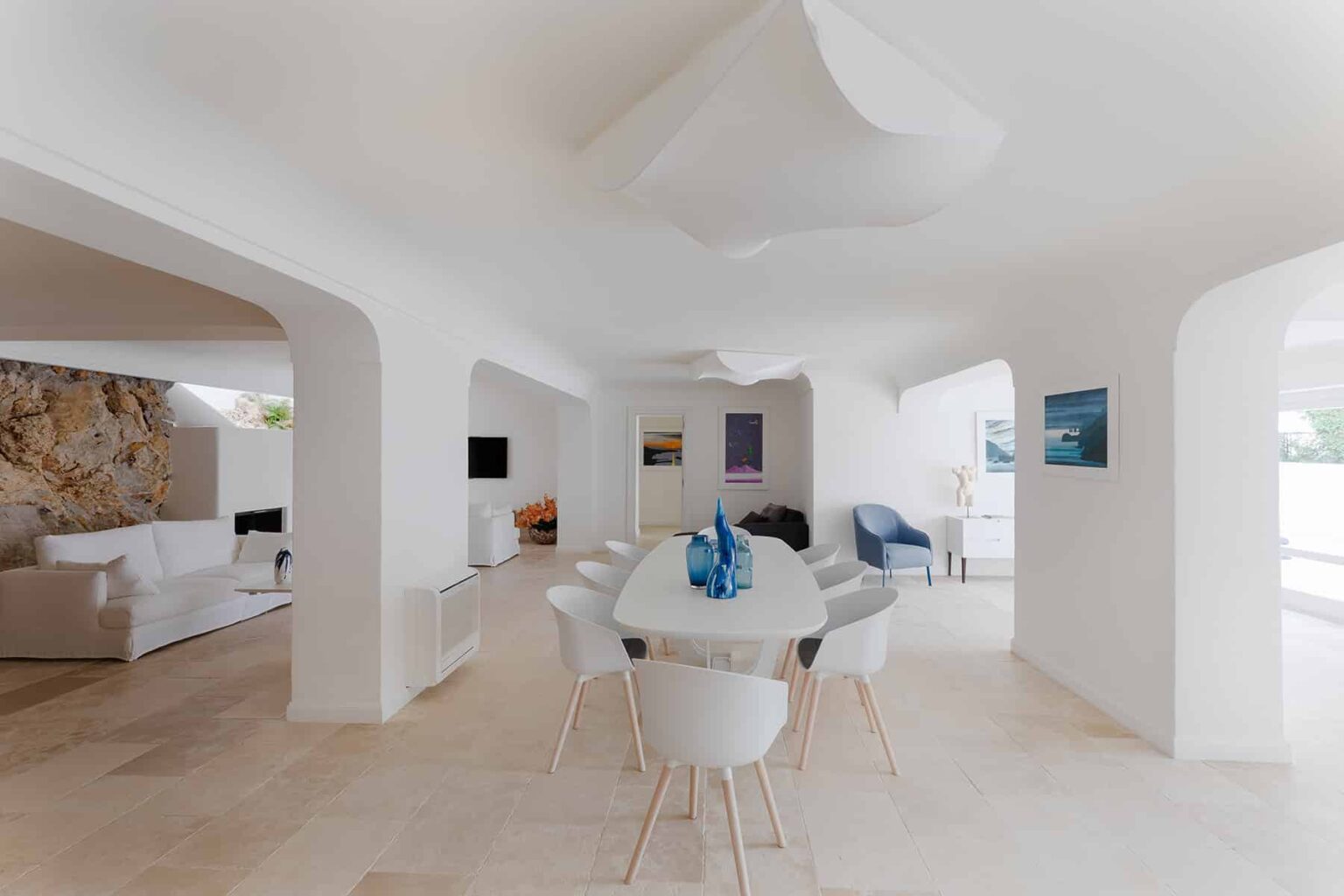 Modern open-concept dining area featuring a white table with blue decor, surrounded by stylish chairs, and bright natural light illuminating the space through large windows. The room showcases a minimalist design with smooth curved architectural elements and neutral-toned flooring. | Only on SopranoVillas