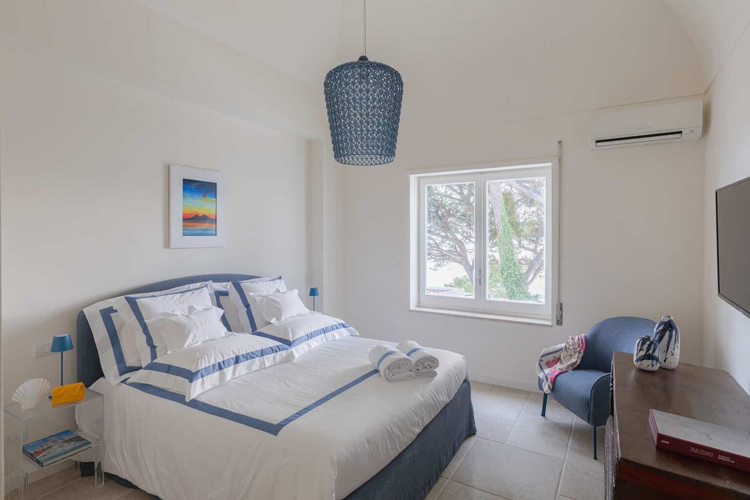 Cozy bedroom featuring a double bed with white linens and blue accents, a stylish blue pendant light, a window with natural light, and a small reading chair next to a wooden desk. Ideal for relaxation and comfort. | Only on SopranoVillas