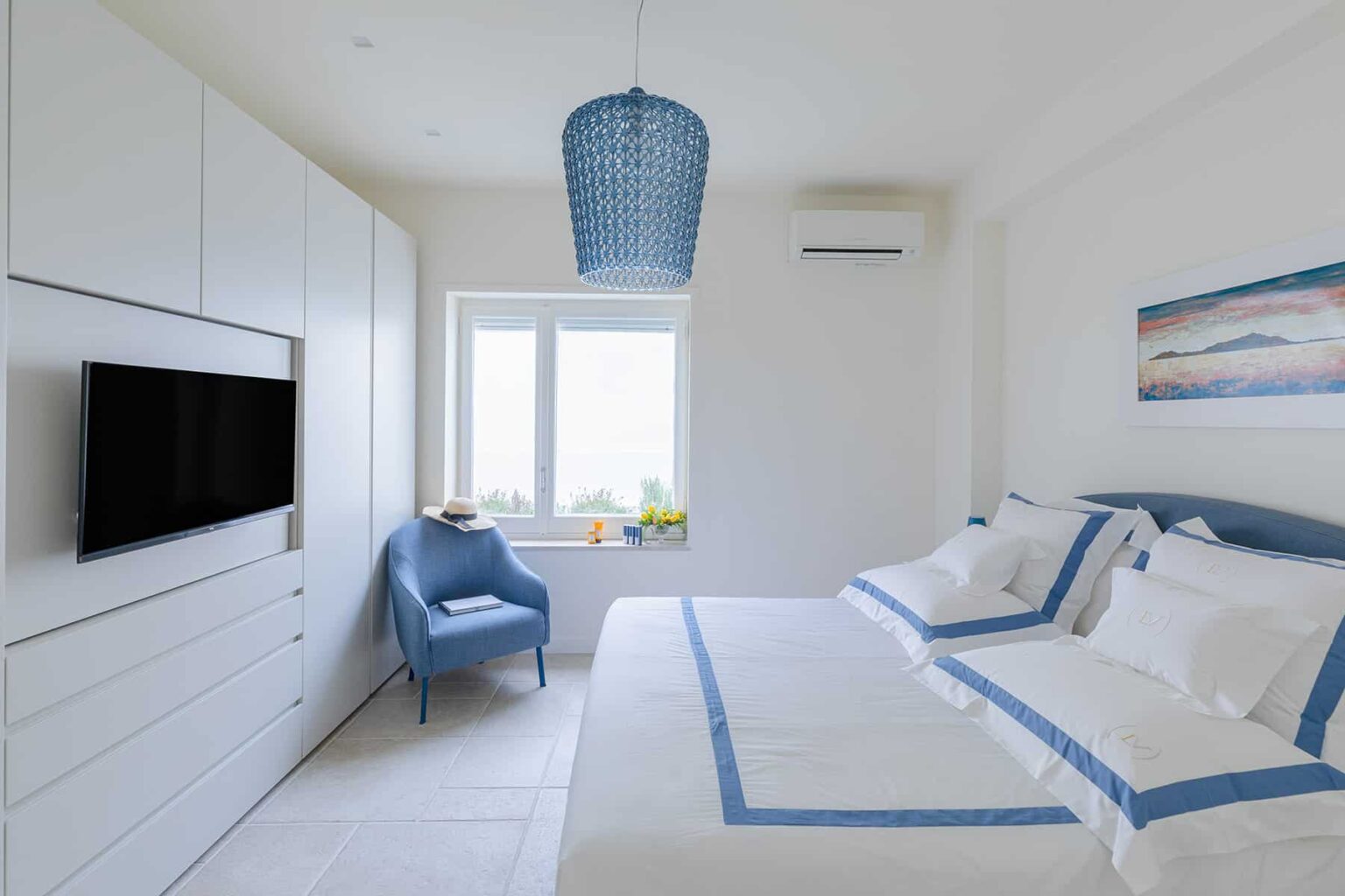 Modern bedroom with blue accents, featuring a cozy bed with white linens and blue trim, a stylish blue chair, wall-mounted TV, and a large window for natural light. | Only on SopranoVillas