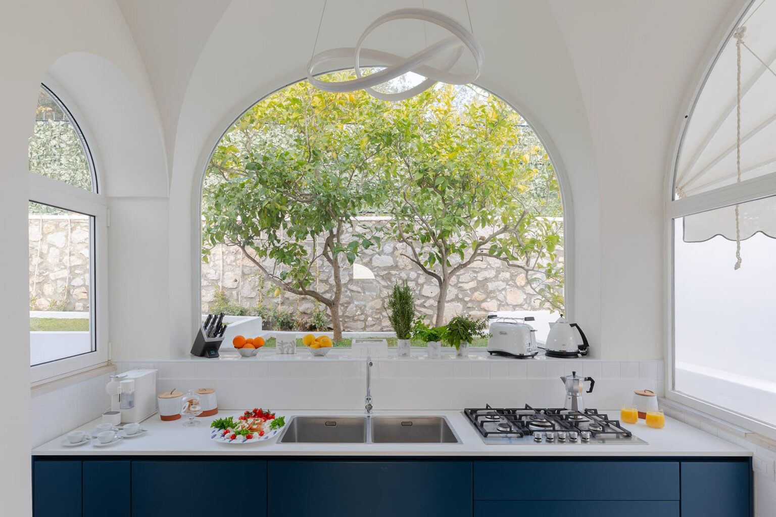 Bright and modern kitchen featuring a blue island, stainless steel gas stove, and fresh ingredients on the counter, with large arched windows showcasing lush greenery outside. | Only on SopranoVillas