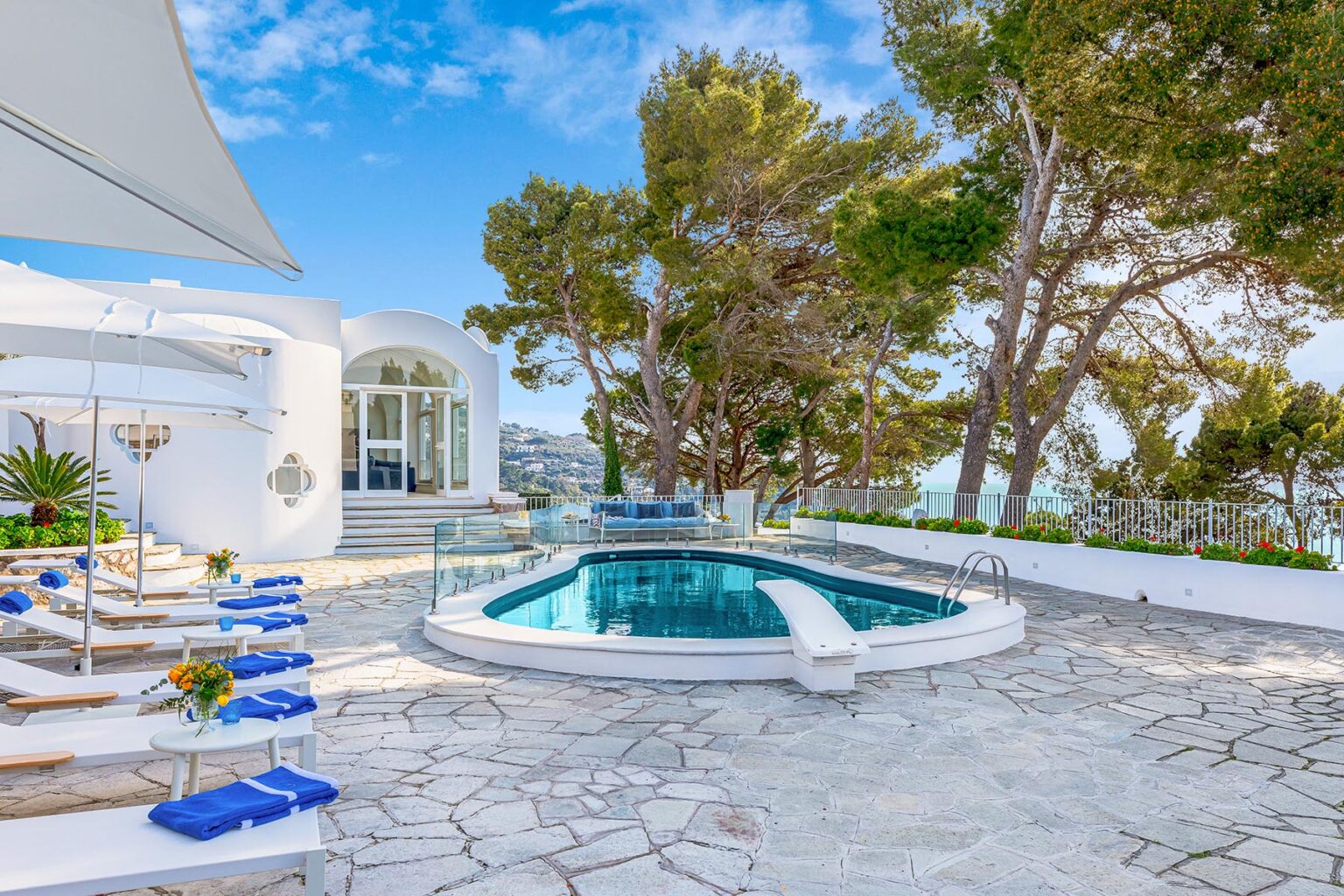 Luxurious villa outdoor area featuring a serene pool, sun loungers with blue cushions, and lush greenery under a clear blue sky. Ideal for relaxation and outdoor gatherings. | Only on SopranoVillas