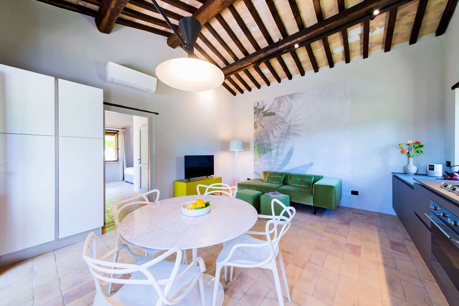 Modern kitchen and dining area featuring a round table with fruit centerpiece, stylish white chairs, a green sofa, and exposed wooden beams on the ceiling. Bright and inviting interior design with natural light. | Only on SopranoVillas