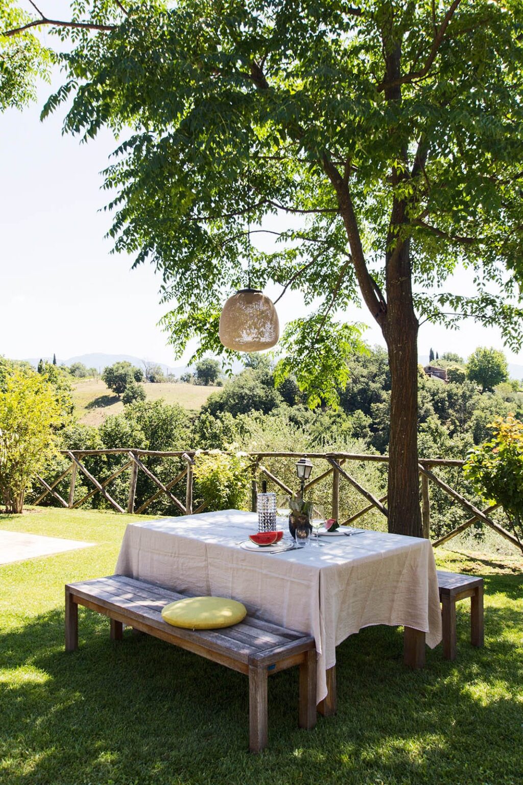 Outdoor dining setup under a tree with a hanging light fixture, featuring a table set with drinks and a scenic view of lush greenery and rolling hills in the background. Ideal for summer gatherings and al fresco dining experiences. | Only on SopranoVillas
