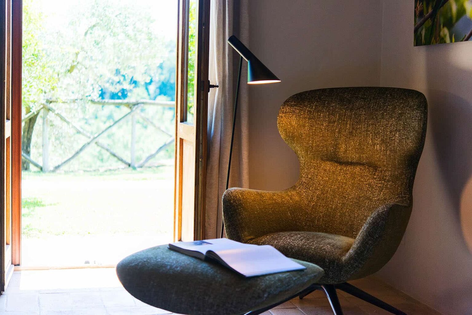 Cozy reading nook featuring a stylish green armchair and ottoman, illuminated by a modern lamp, with an open book resting on the seat and large windows offering a view of the outdoors. | Only on SopranoVillas