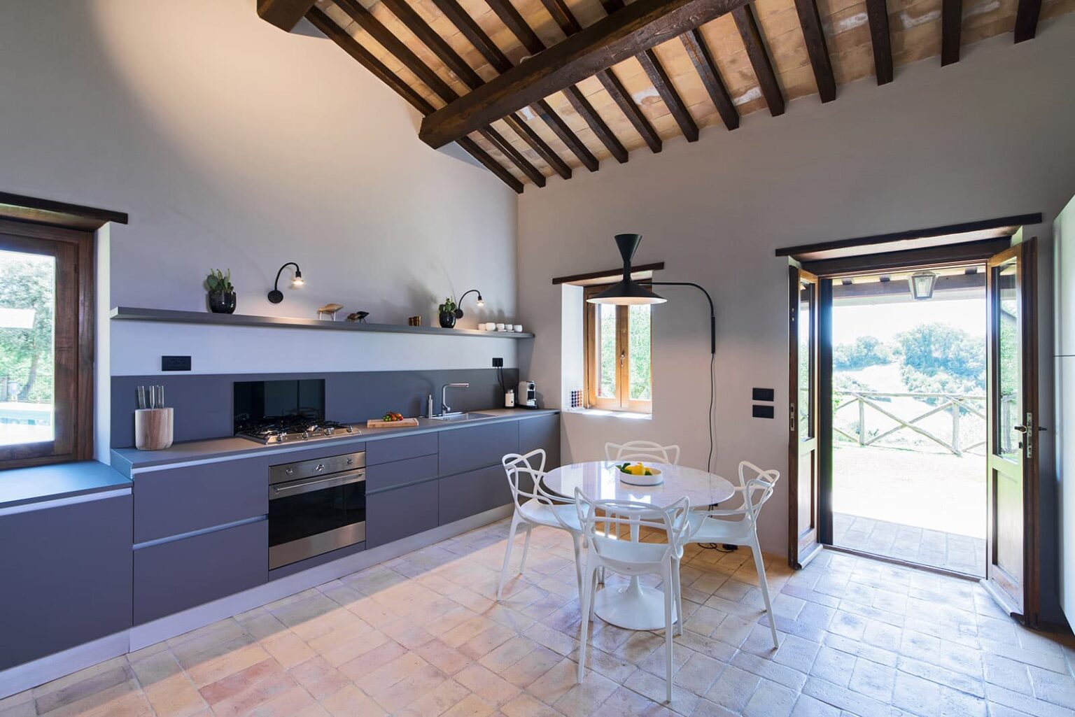 Modern kitchen with wooden beams, featuring sleek gray cabinetry, stainless steel appliances, and a dining table with white chairs, illuminated by natural light from an open door leading to a scenic outdoor view. | Only on SopranoVillas