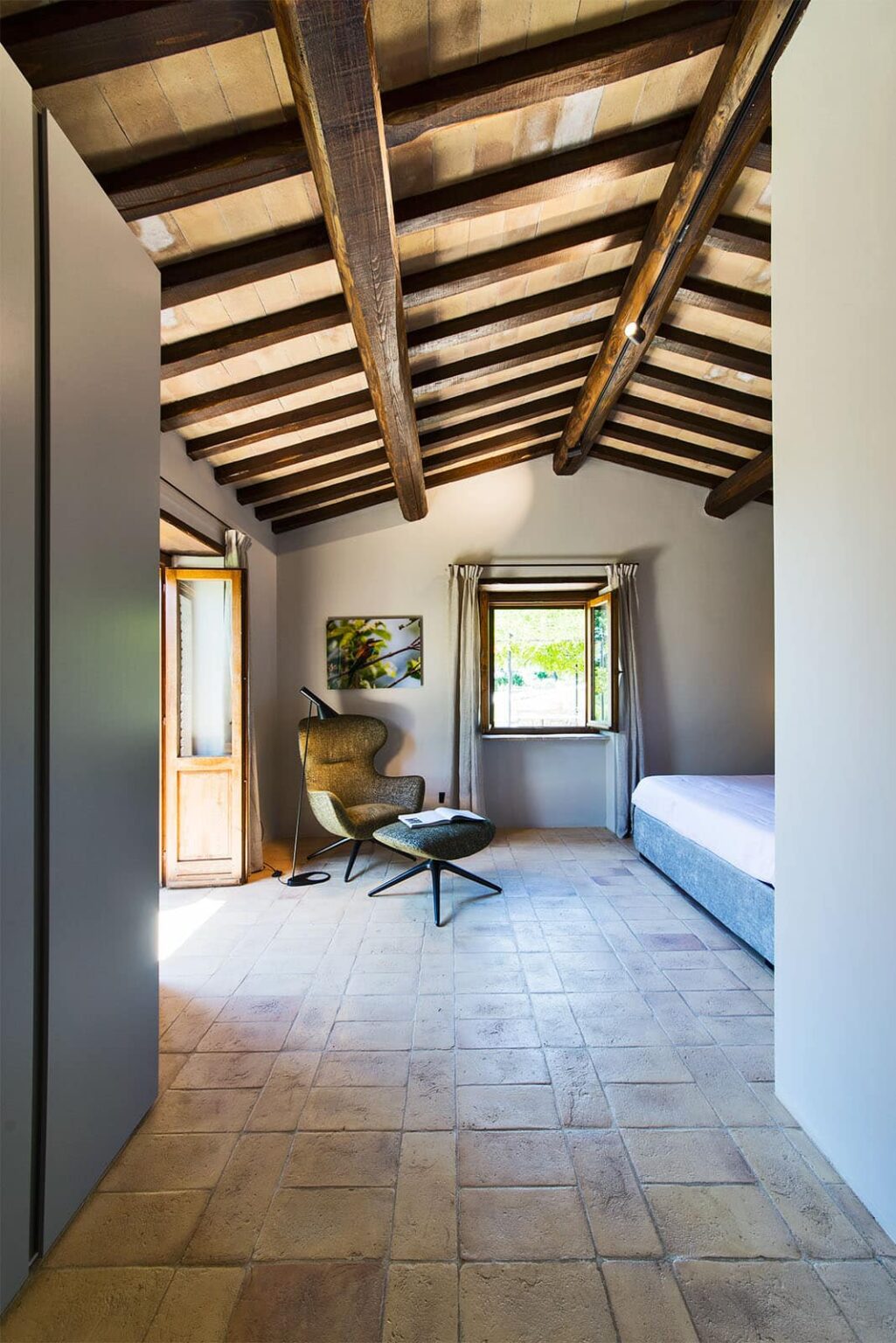 Cozy bedroom with wooden beam ceiling, featuring a comfortable chair and small coffee table, large window with natural light, and a minimalist decor style. | Only on SopranoVillas