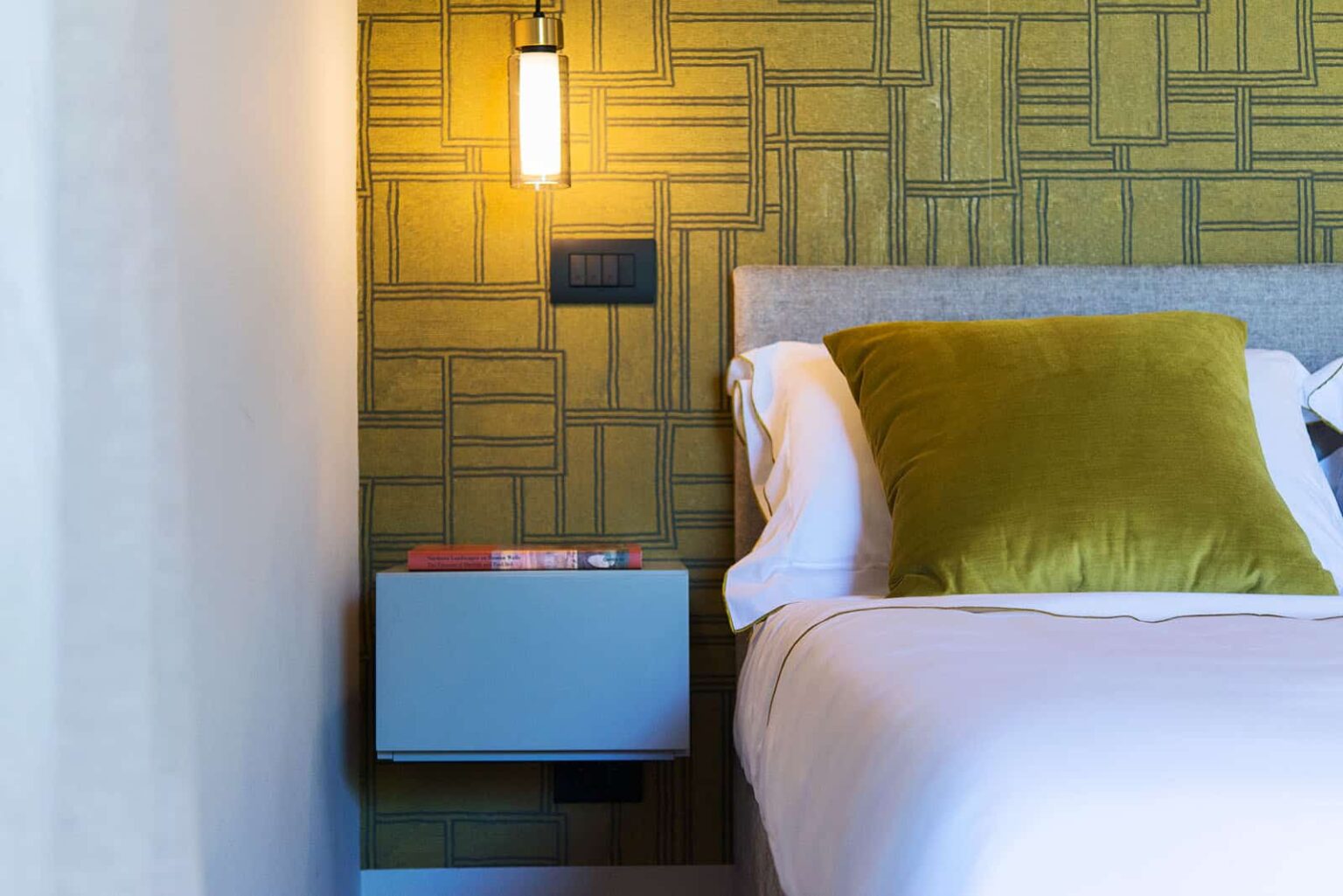 Modern bedroom interior featuring a plush bed with decorative pillows, a sleek nightstand with books, and a stylish wall lamp illuminating a textured yellow-green wallpaper. | Only on SopranoVillas