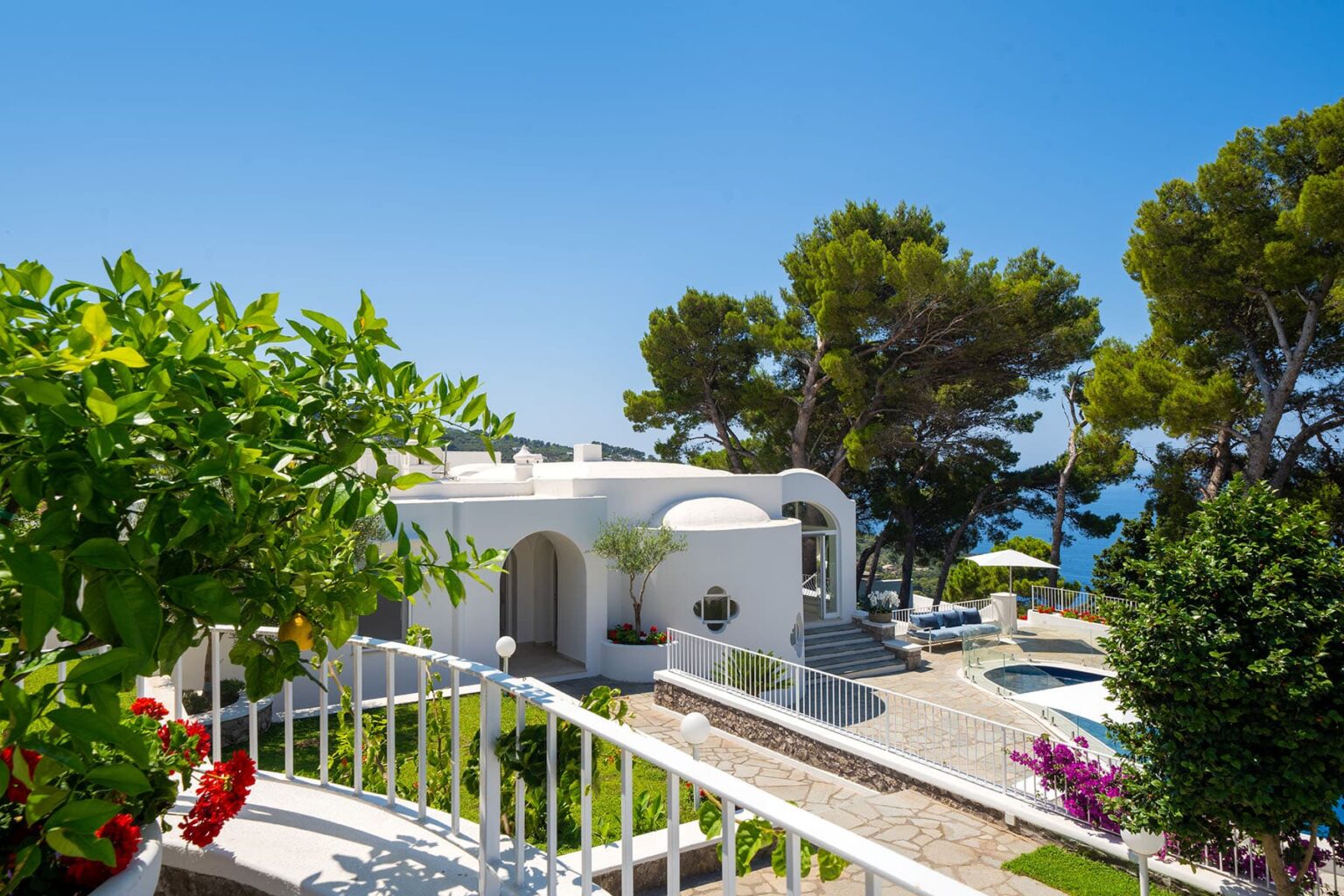 Stunning coastal villa surrounded by lush greenery and vibrant flowers, featuring a pool and panoramic views of the sea under a clear blue sky. Ideal for luxury vacations and relaxation. | Only on SopranoVillas