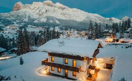 Snow-covered chalet in the Dolomites with warm lights glowing, surrounded by pine trees and majestic mountains at dusk.