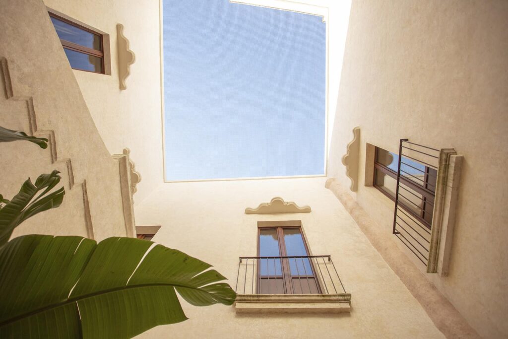 View looking up from a courtyard, showcasing a clear blue sky framed by architectural elements and windows of a building, with a green plant in the foreground. | Only on SopranoVillas