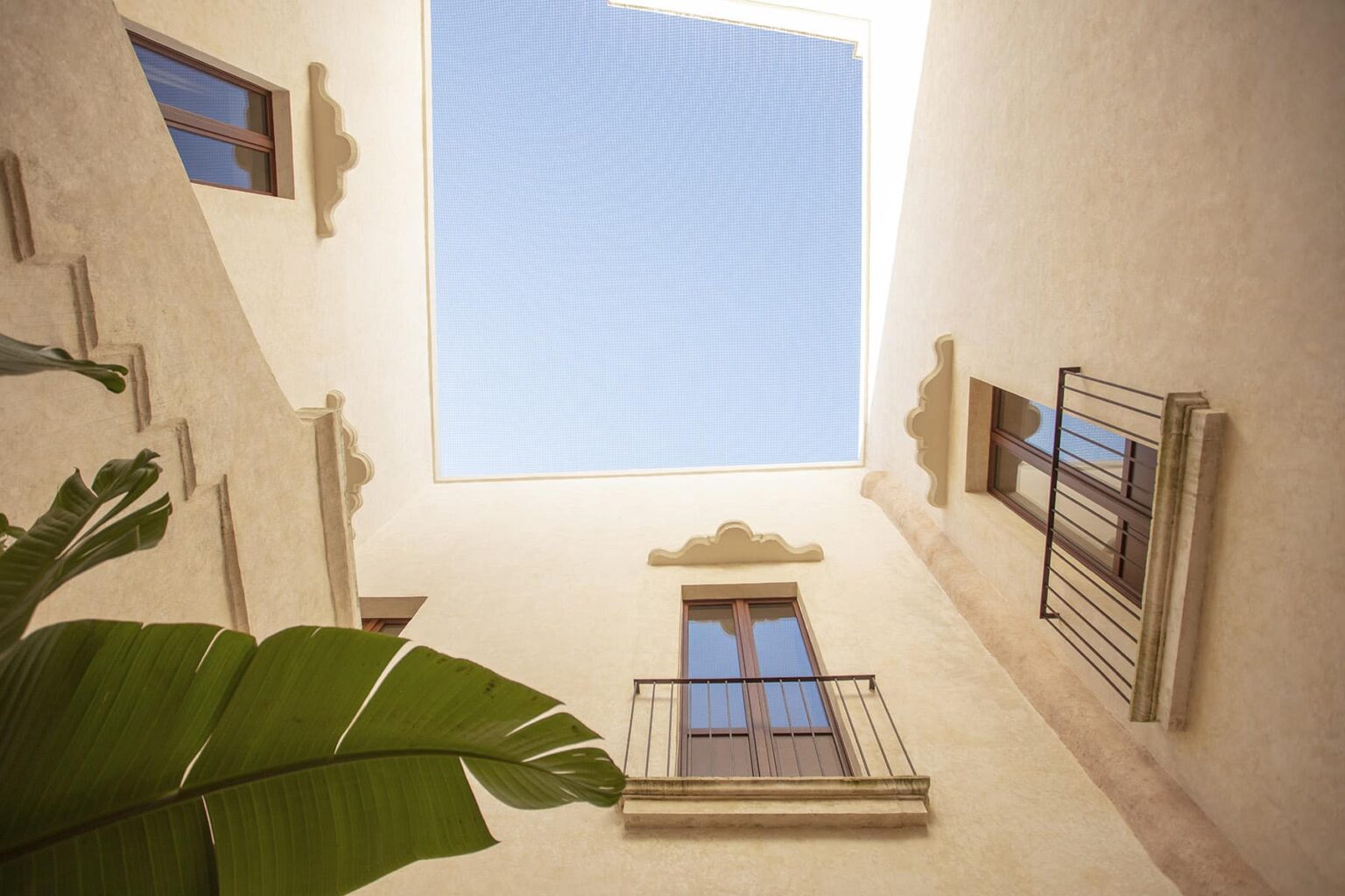 View looking up from a courtyard, showcasing a clear blue sky framed by architectural elements and windows of a building, with a green plant in the foreground. | Only on SopranoVillas
