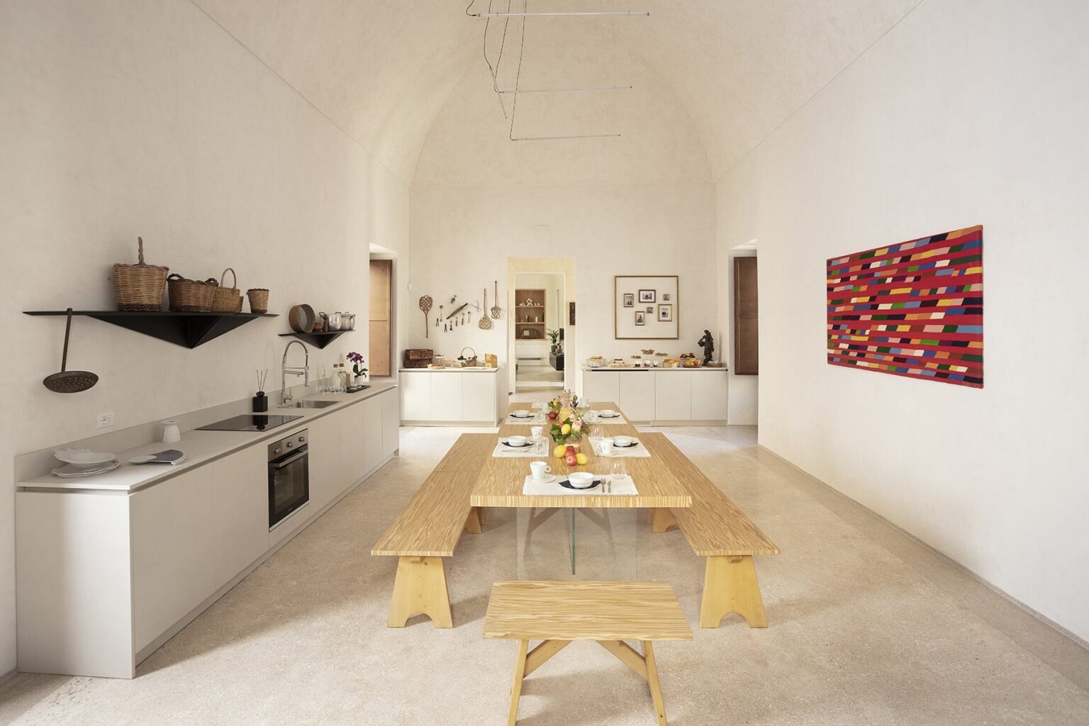 Modern minimalist kitchen and dining area with a large wooden table, white walls, and colorful artwork. The kitchen features sleek cabinetry and a contemporary oven, while natural light floods the space, enhancing its airy feel. | Only on SopranoVillas