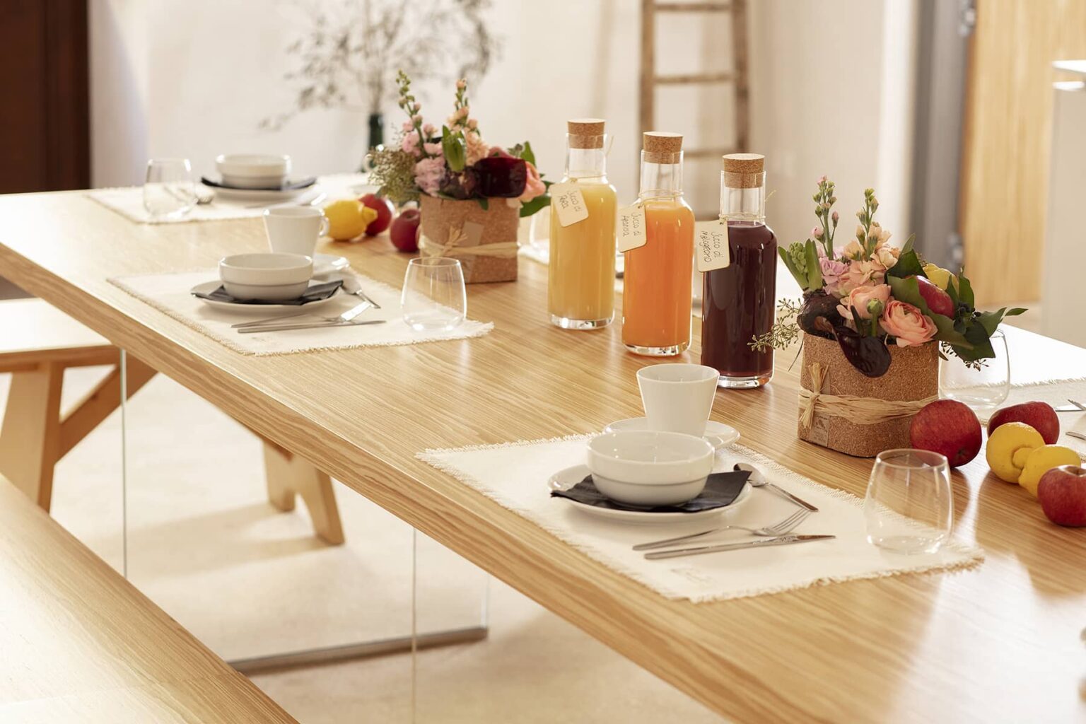 A beautifully set dining table with wooden accents, featuring white dishware, glasses, and colorful drinks in glass bottles, complemented by floral arrangements and fresh fruits, creating an inviting atmosphere for a meal or gathering. | Only on SopranoVillas
