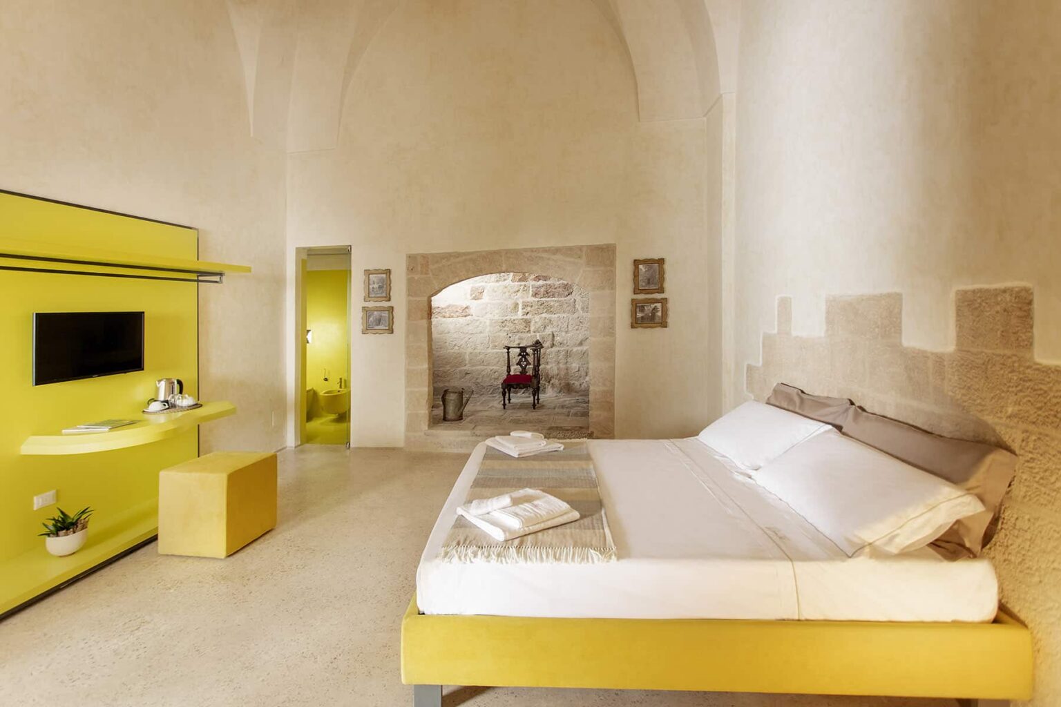 Modern bedroom interior featuring a spacious layout with a yellow accent wall, minimalist furniture, and natural light. The room includes a double bed, a stylish television unit, and decorative elements that enhance the contemporary aesthetic. | Only on SopranoVillas