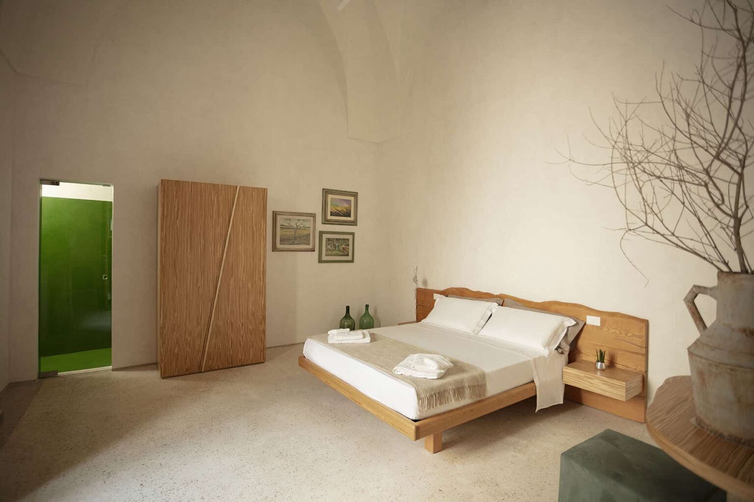 Modern minimalist bedroom featuring a wooden bed with white linens, a natural wood wardrobe, and decorative artwork on the walls. A green door adds a pop of color to the serene, neutral-toned space. | Only on SopranoVillas