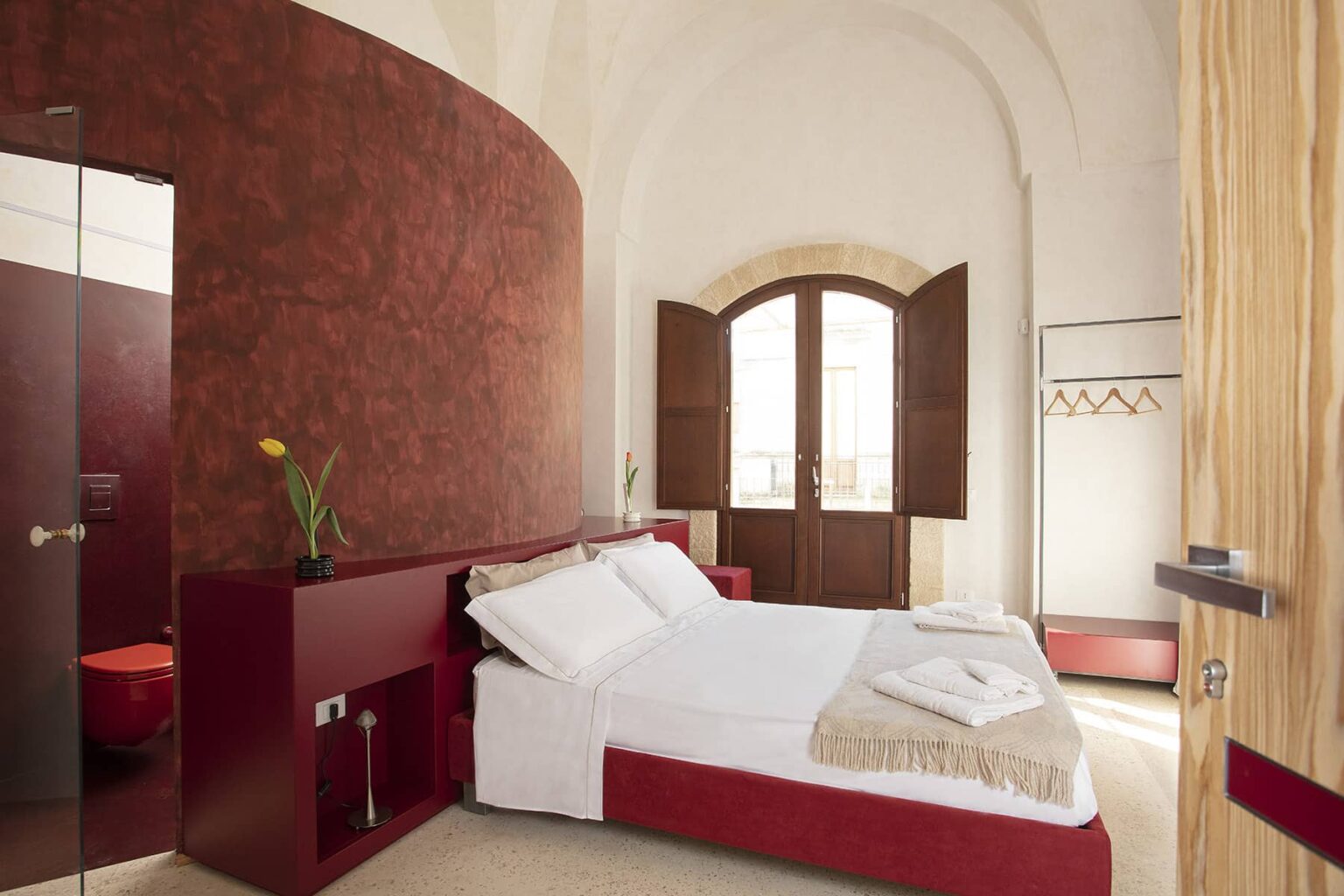 Modern hotel room featuring a comfortable double bed with white linens, elegant red walls, large wooden windows, and minimalist decor. Ideal for travelers seeking a stylish and cozy accommodation. | Only on SopranoVillas
