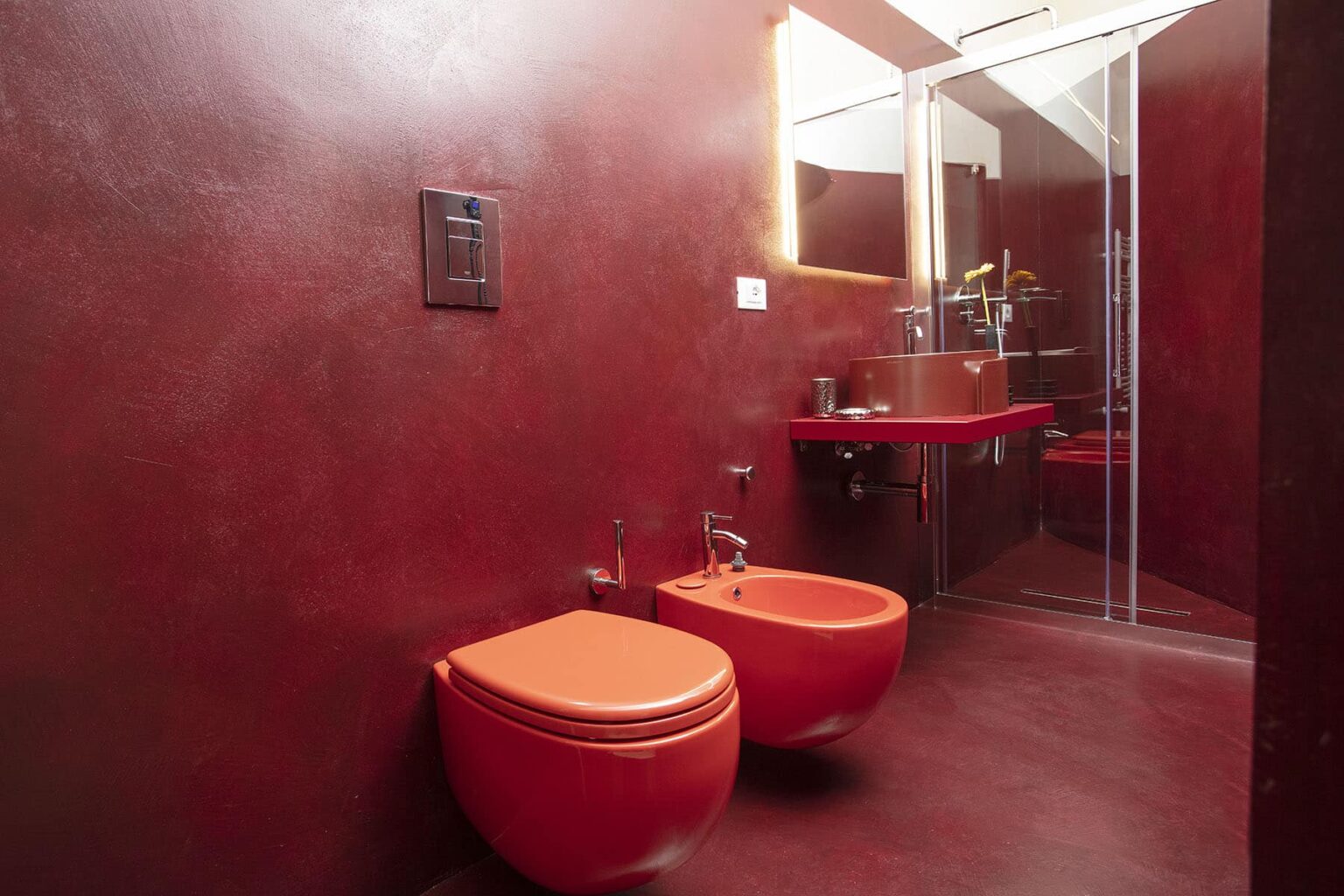 Modern bathroom featuring vibrant red walls, stylish orange bidets, and a glass shower enclosure. The design emphasizes a contemporary aesthetic with sleek fixtures and ample natural light. | Only on SopranoVillas