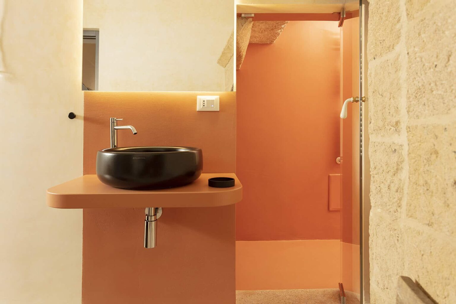 Modern bathroom featuring a stylish black sink on a floating shelf, with an orange accent wall and a stone-textured shower area. Ideal for contemporary interior design inspiration. | Only on SopranoVillas
