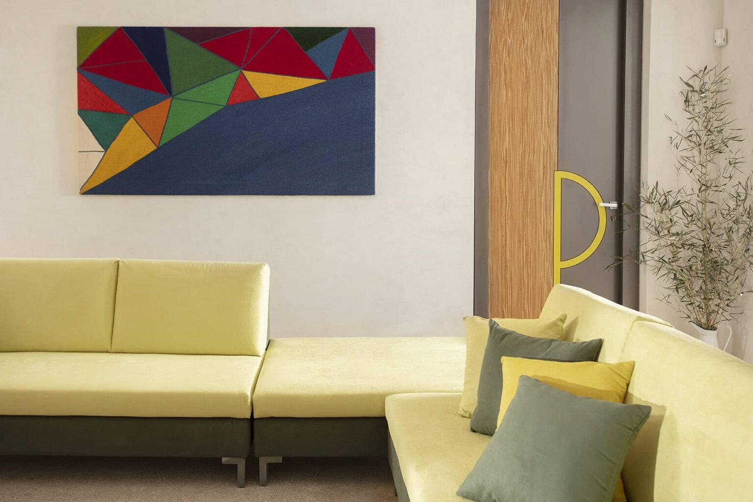 Modern living room featuring a yellow sectional sofa adorned with colorful cushions, complemented by an abstract geometric painting on the wall. The space is bright and inviting, showcasing contemporary design elements. | Only on SopranoVillas