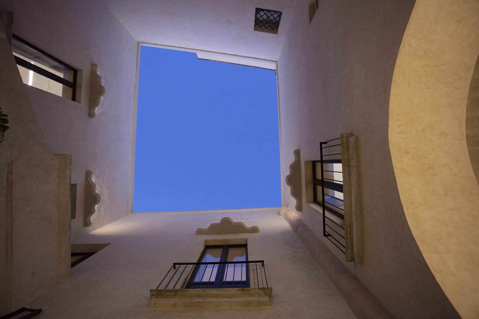 Looking up at a clear blue sky from an enclosed courtyard with white walls and balconies, showcasing architectural details and a serene atmosphere. | Only on SopranoVillas