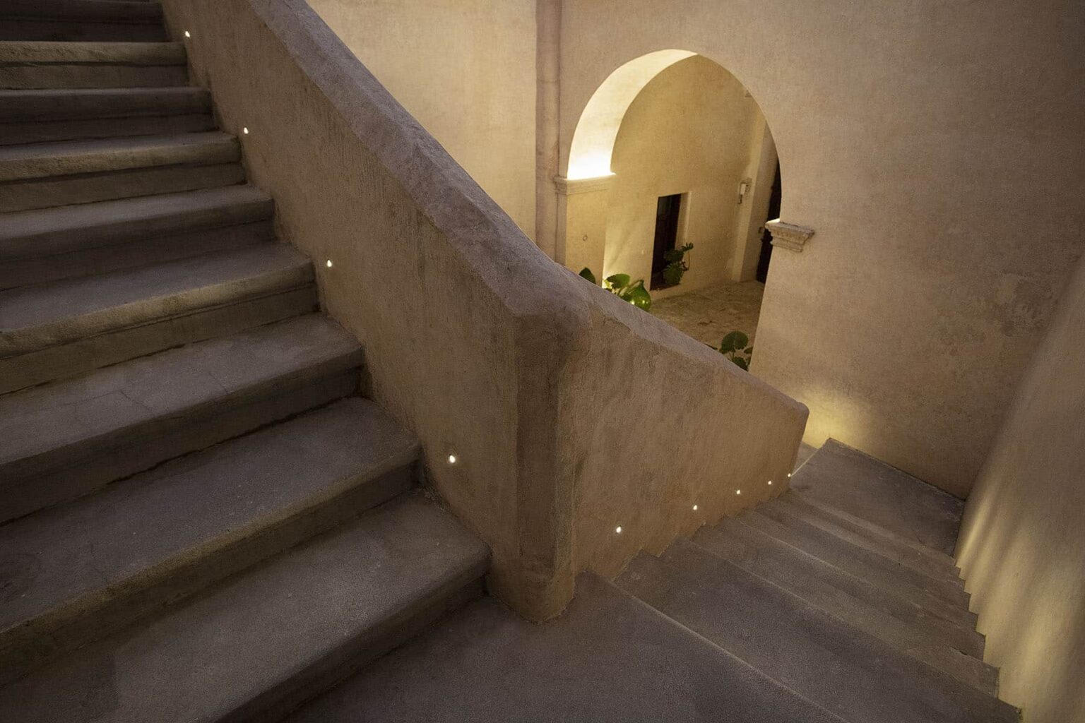 Interior view of a beautifully designed staircase with soft lighting, leading to an arched doorway. The staircase features smooth concrete steps and a minimalist aesthetic, surrounded by warm, inviting tones. | Only on SopranoVillas