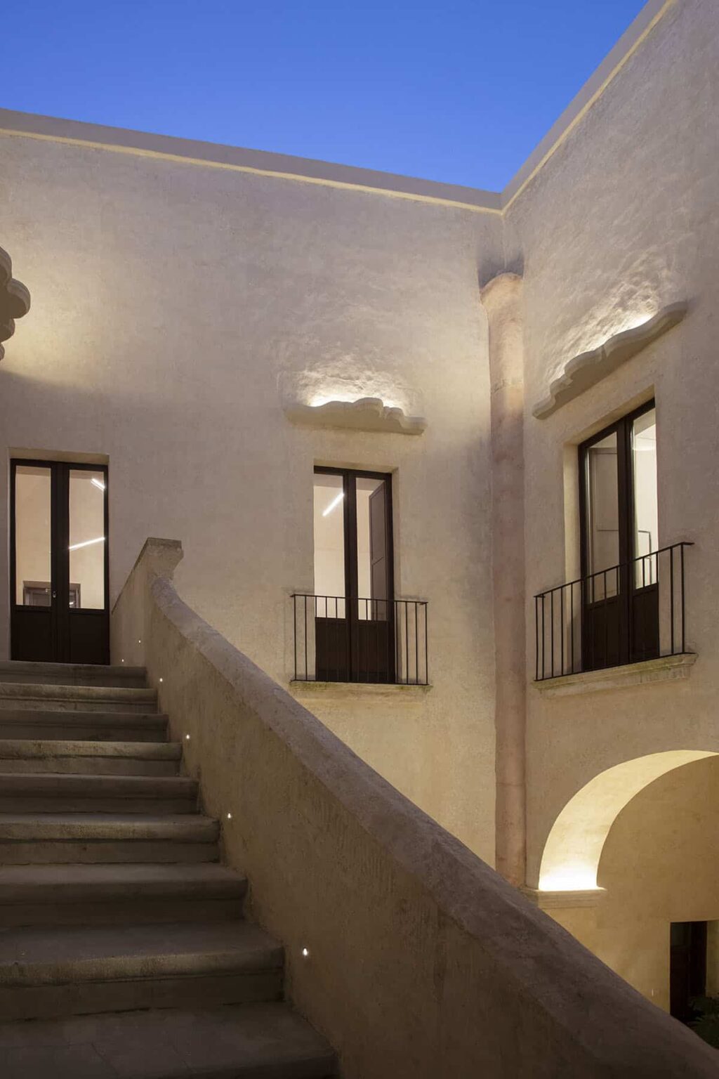 Interior view of a modern building featuring a staircase, large windows with black railings, and a softly illuminated wall design, showcasing contemporary architectural elements. | Only on SopranoVillas