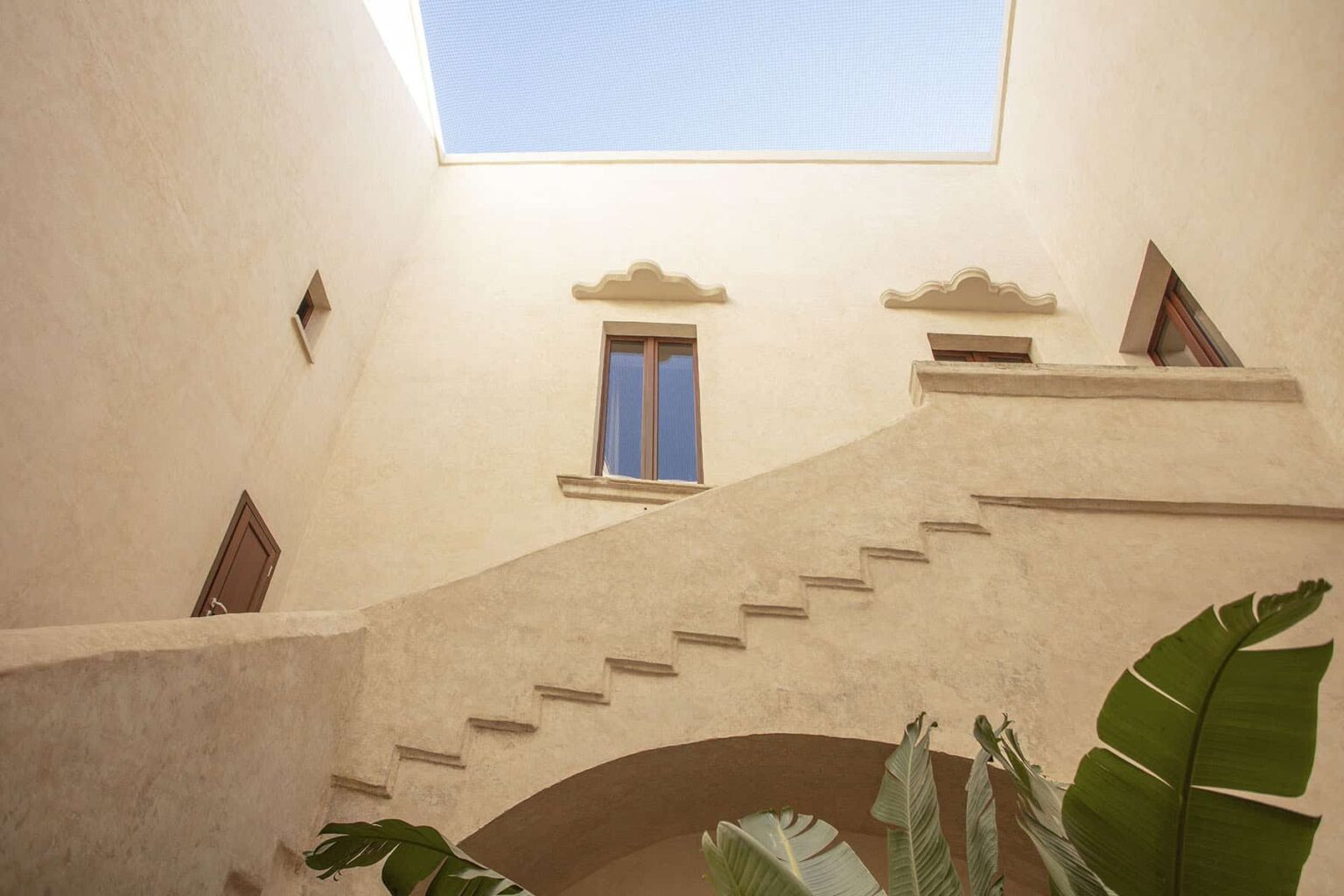 Interior view of a traditional architectural space featuring a staircase, large windows, and natural light streaming from an overhead opening, with green foliage in the foreground. | Only on SopranoVillas