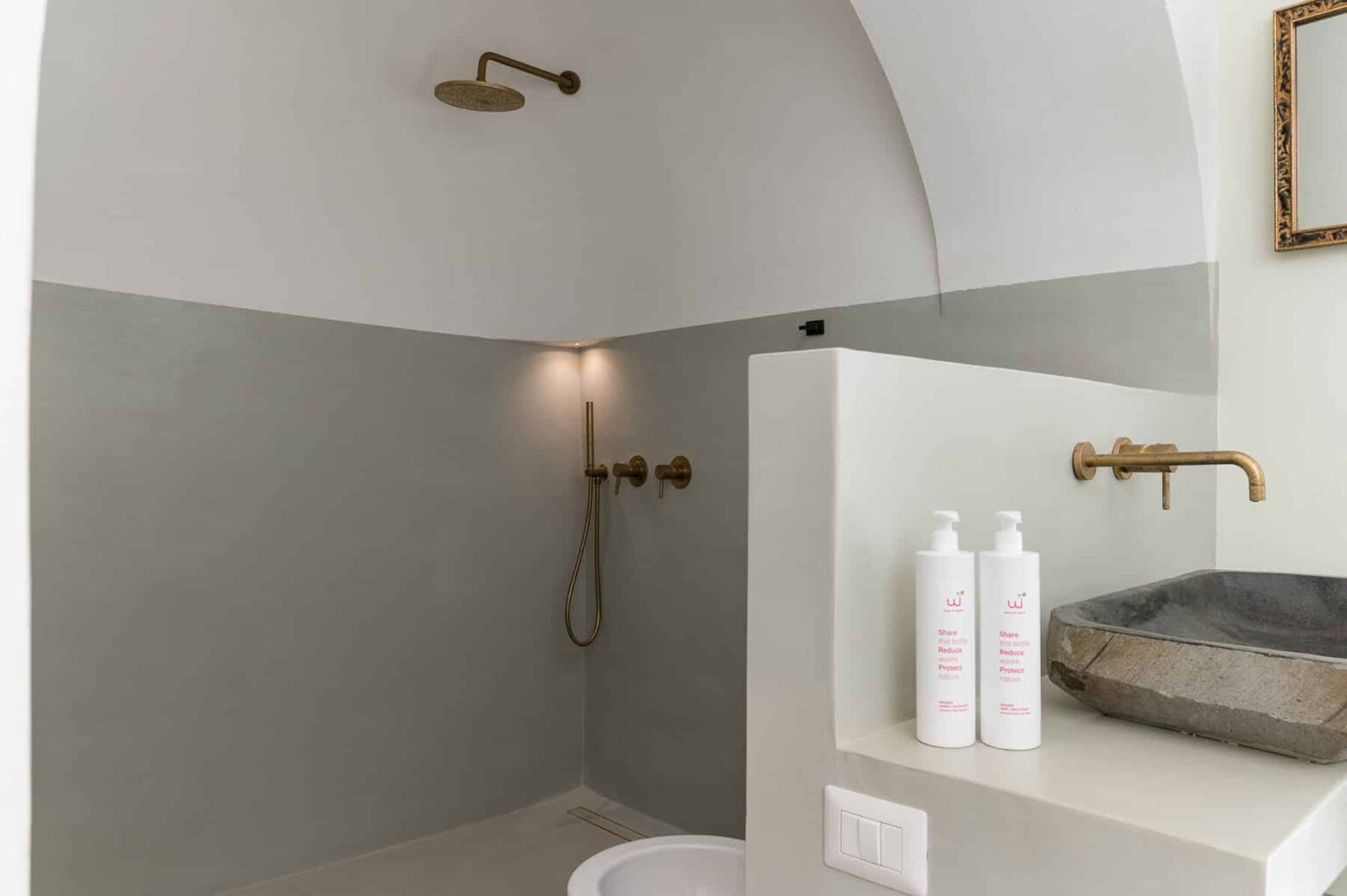 Modern bathroom design featuring a minimalist shower with a curved ceiling, elegant brass fixtures, and a stylish stone sink. Three white shampoo bottles are displayed on a shelf, enhancing the contemporary aesthetic. | Only on SopranoVillas