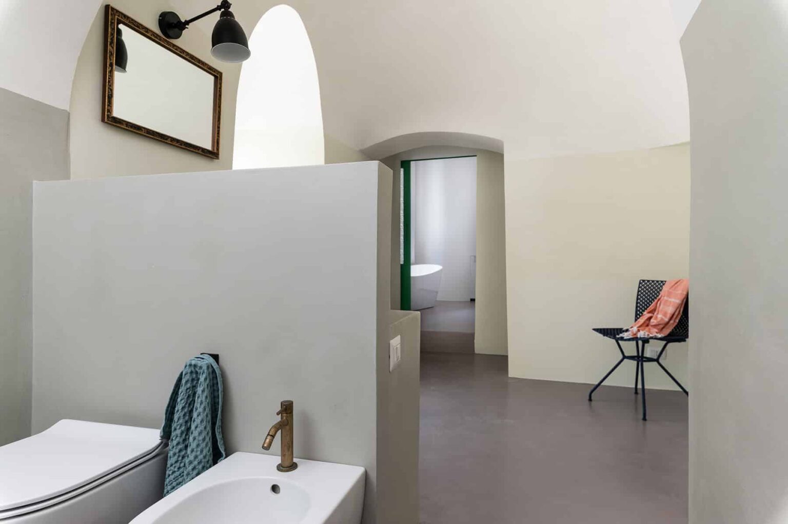 Modern bathroom interior featuring minimalist design, a gray partition, sleek fixtures, and a stylish chair. Natural light filters through an arched doorway, enhancing the spacious feel. | Only on SopranoVillas