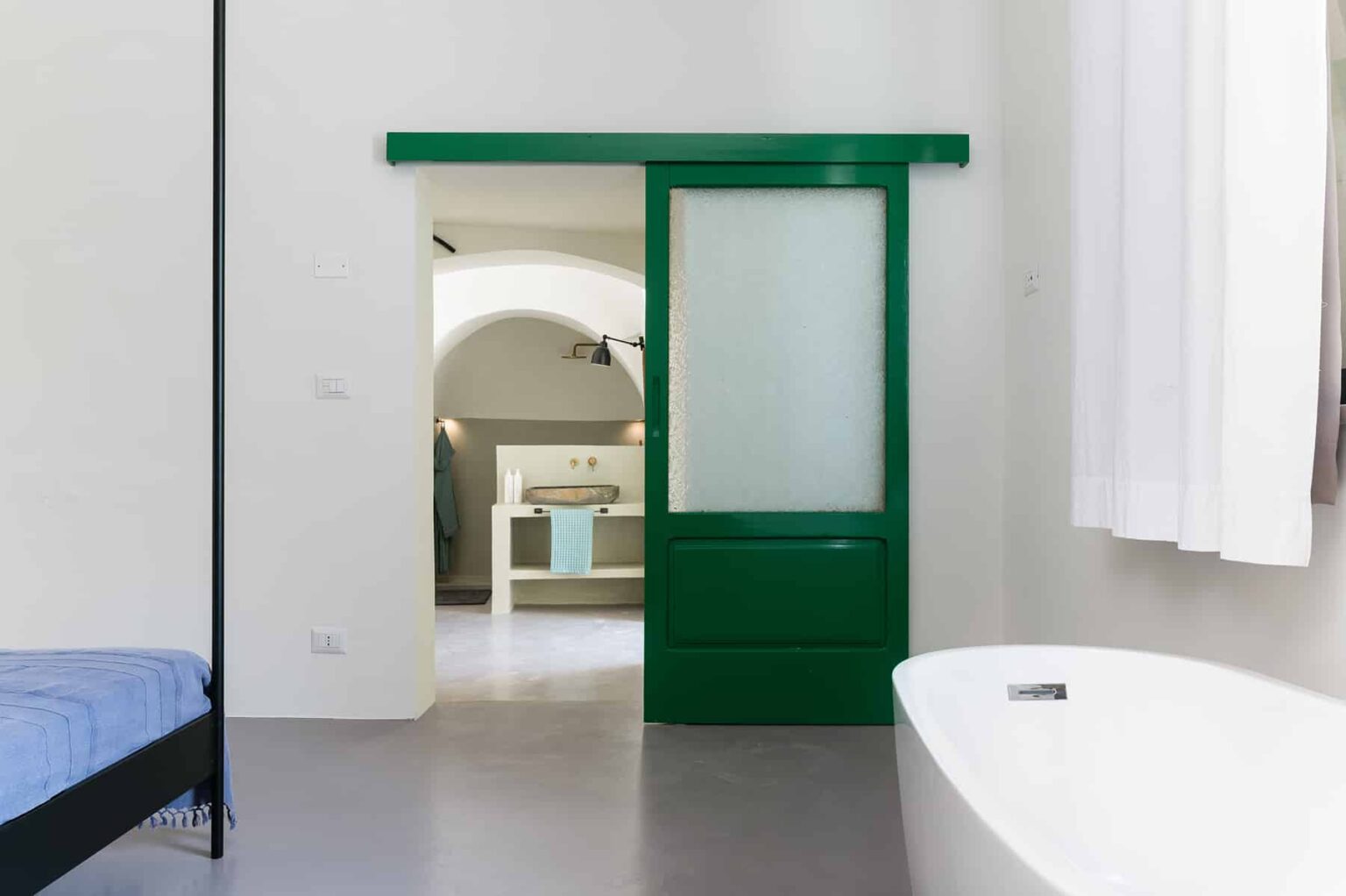 Modern interior design featuring a green sliding door leading to a stylish bathroom. The open space includes a freestanding bathtub, minimalistic decor, and natural light streaming through sheer curtains, creating a serene atmosphere. | Only on SopranoVillas