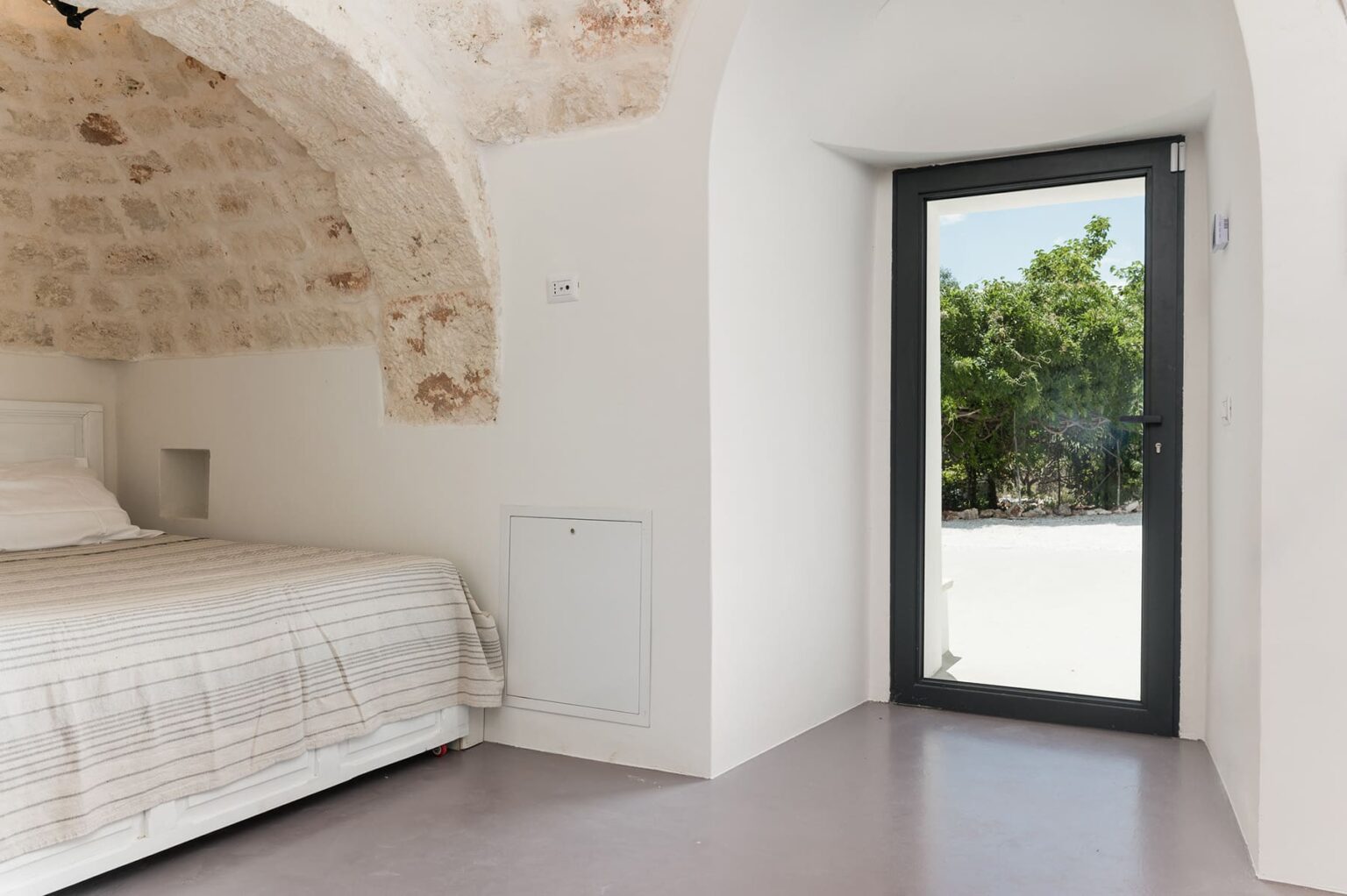 Modern bedroom with a stone arch ceiling, featuring a bed with light bedding and a large glass door leading to an outdoor view. | Only on SopranoVillas