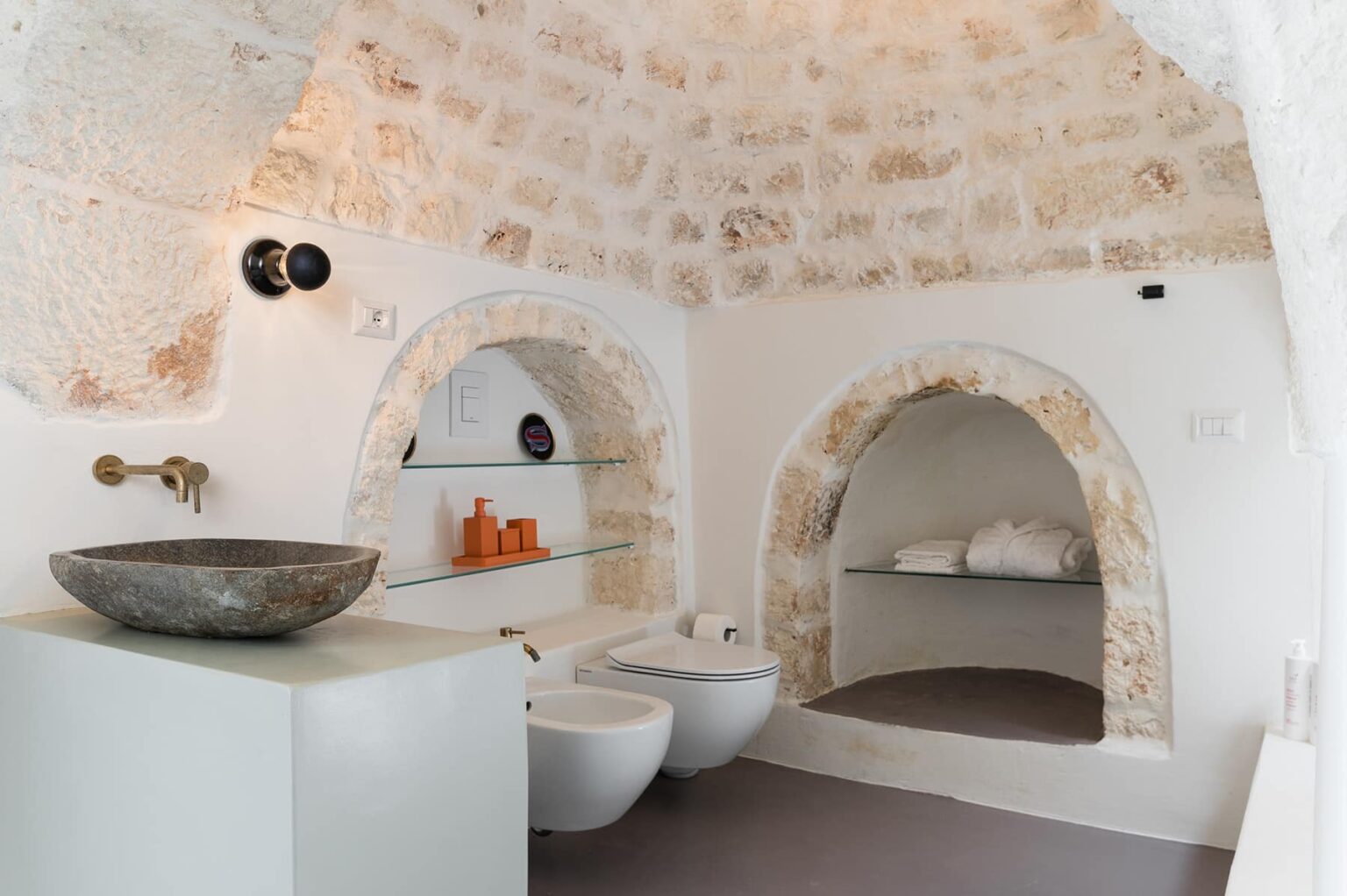Modern bathroom design with arched stone walls, featuring a sleek sink, bidet, and minimalist shelving. The space is bright and airy, enhanced by natural stone textures and contemporary fixtures. | Only on SopranoVillas