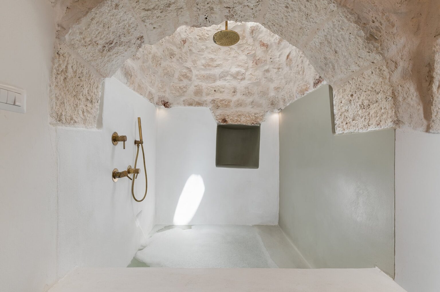 Minimalist bathroom interior featuring a stone arch ceiling, white walls, and modern fixtures including a showerhead and faucet. Natural light illuminates the space, enhancing the serene and spacious atmosphere. | Only on SopranoVillas