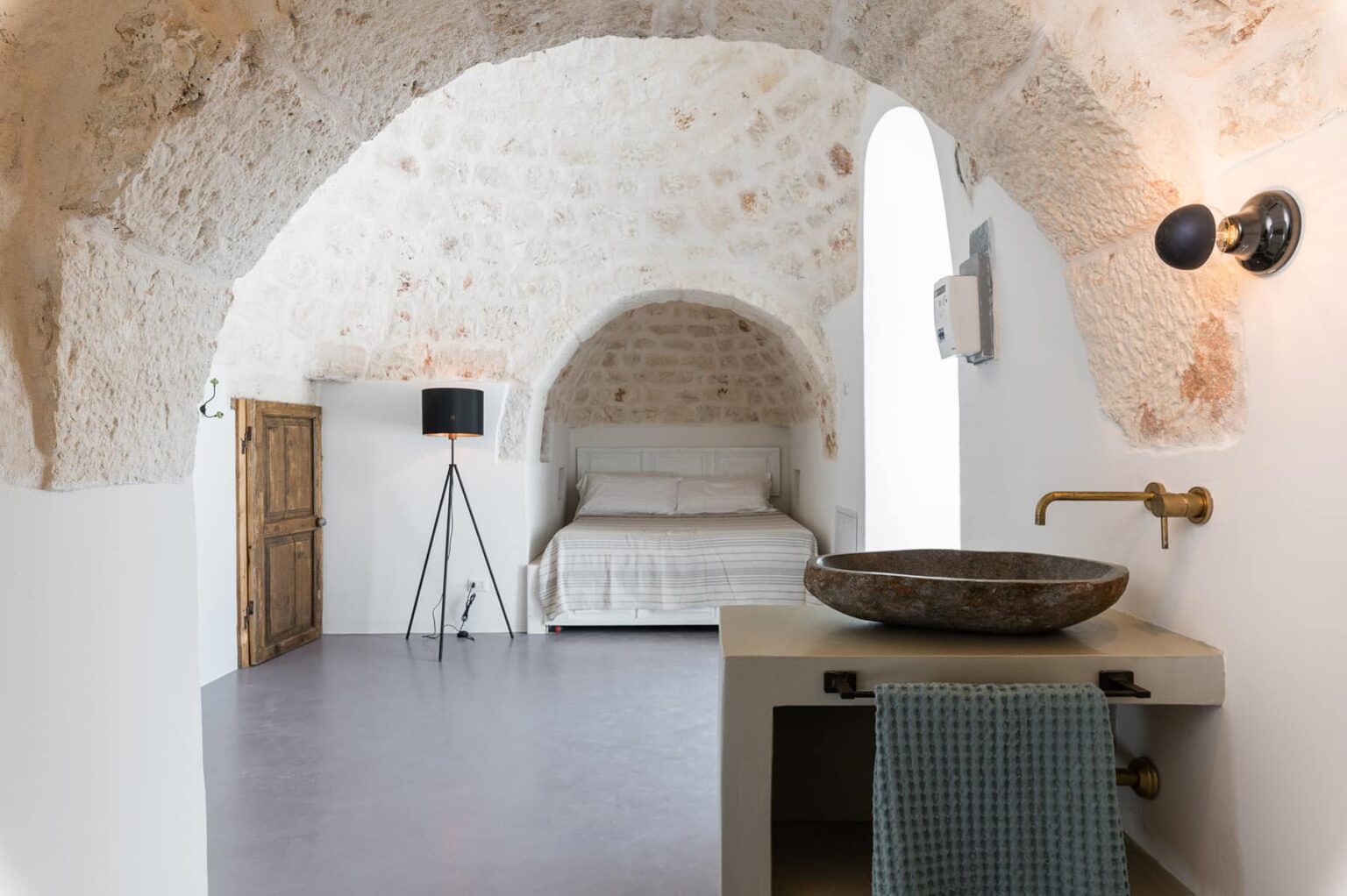 Interior view of a stylish bedroom featuring arched stone ceilings, a minimalist bed, a modern floor lamp, and a rustic sink with a wooden bowl, creating a cozy and elegant atmosphere. | Only on SopranoVillas