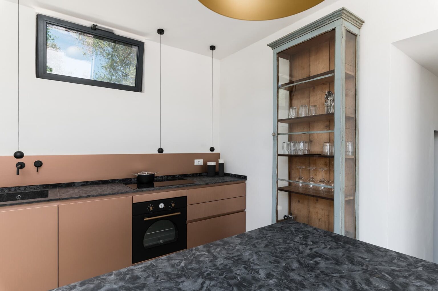 Modern kitchen interior featuring sleek cabinetry in muted tones, a black countertop, built-in oven, and a glass-fronted cabinet displaying glassware, illuminated by natural light from a window. | Only on SopranoVillas