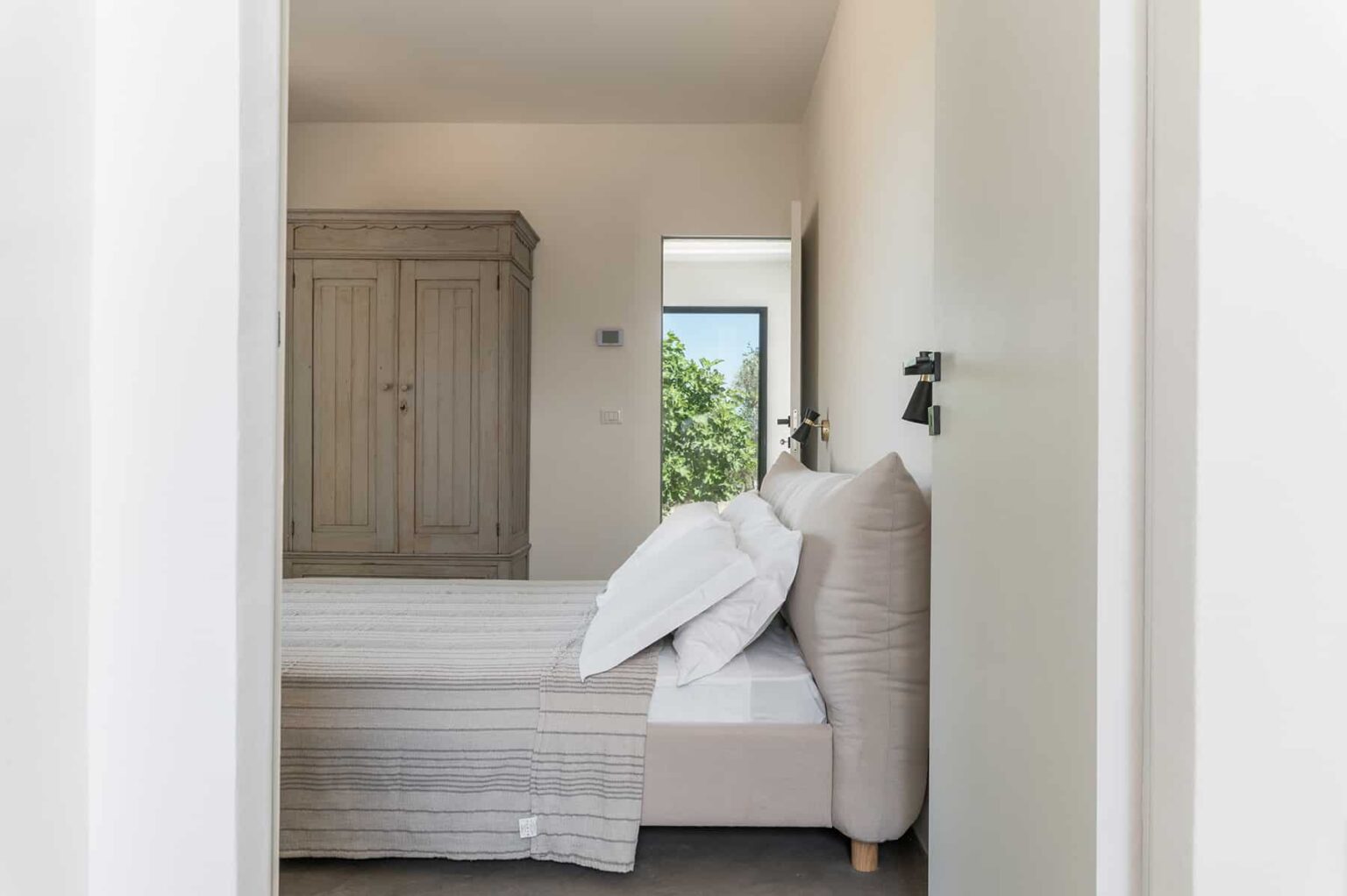 Cozy bedroom interior featuring a bed with white linens and decorative pillows, a wooden wardrobe, and natural light streaming through a window, creating a serene atmosphere. | Only on SopranoVillas