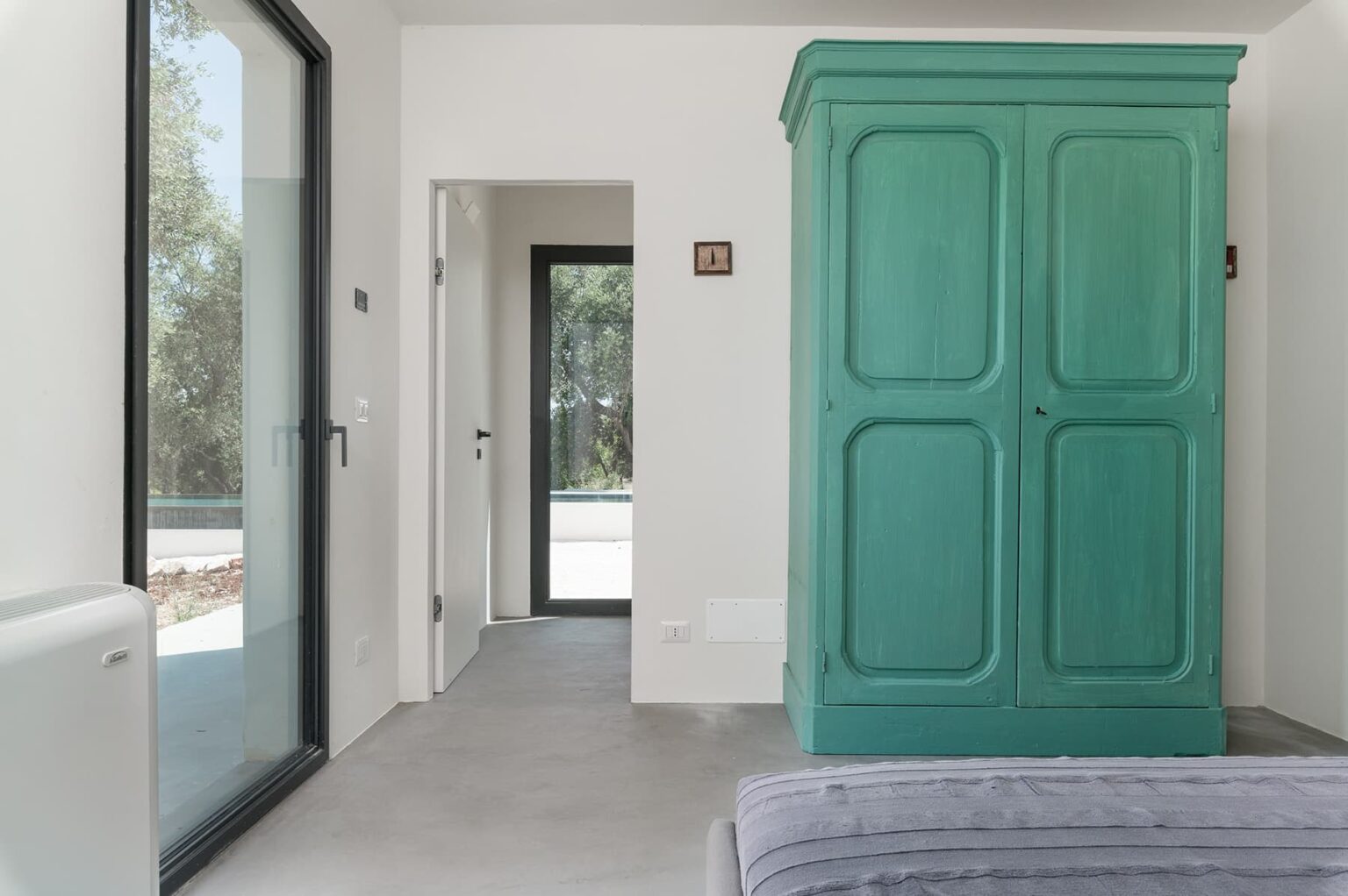Interior view of a modern room featuring a vintage green wardrobe, large windows allowing natural light, and minimalist decor with gray flooring, creating a spacious and inviting atmosphere. | Only on SopranoVillas