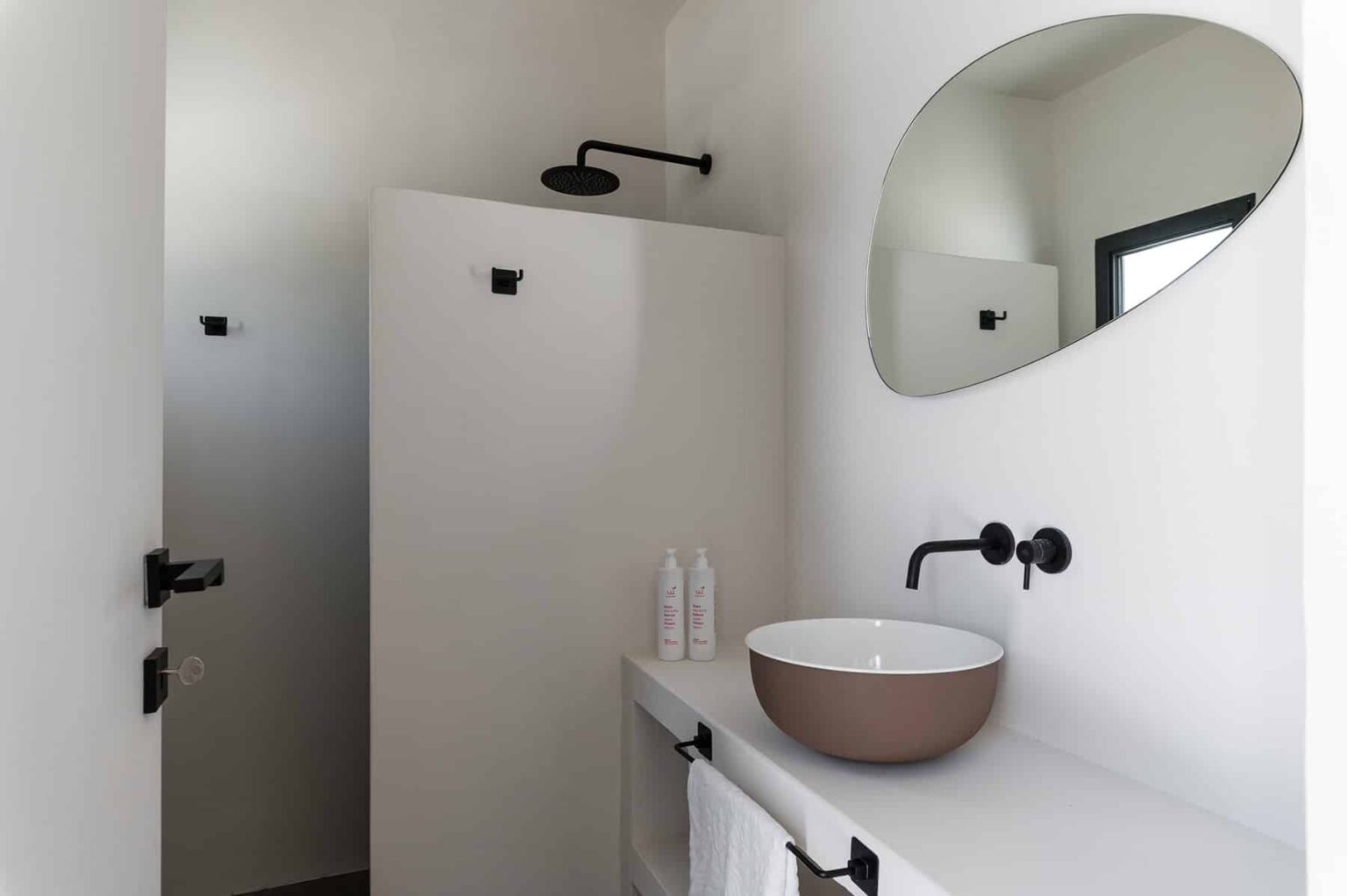 Modern bathroom interior featuring a minimalist design with a round mirror, a brown basin sink, and sleek black fixtures. The white walls and shower area enhance the clean aesthetic, creating a serene and stylish atmosphere. | Only on SopranoVillas