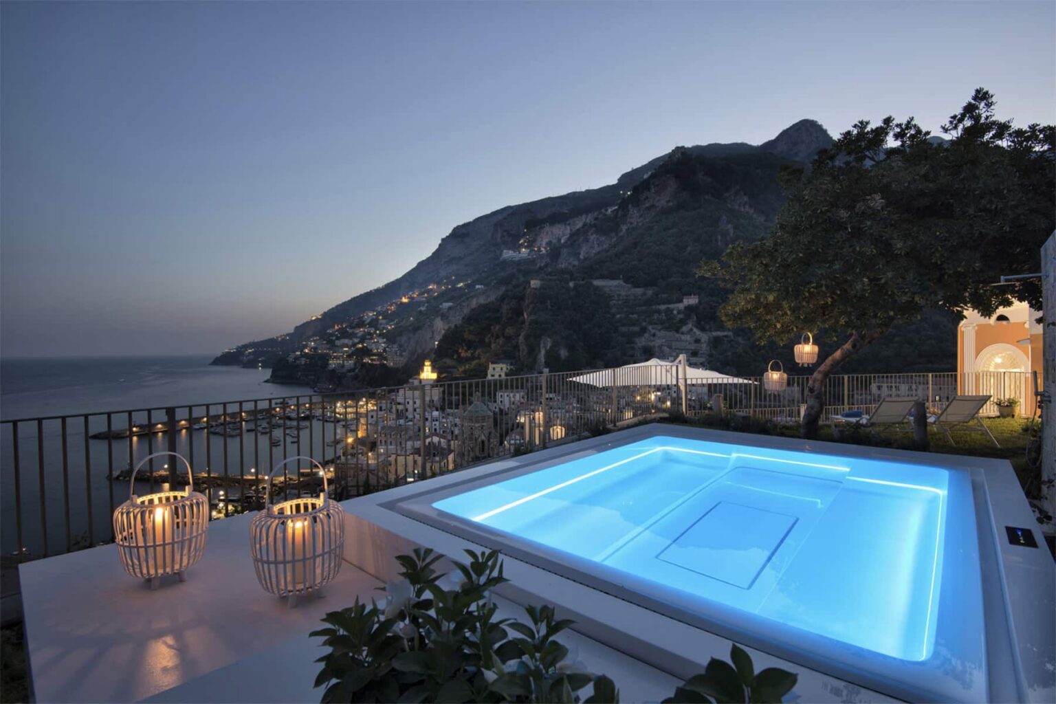 Luxurious outdoor pool with blue lighting, surrounded by lush greenery and lanterns, overlooking a coastal landscape at sunset in a serene setting. | Only on SopranoVillas