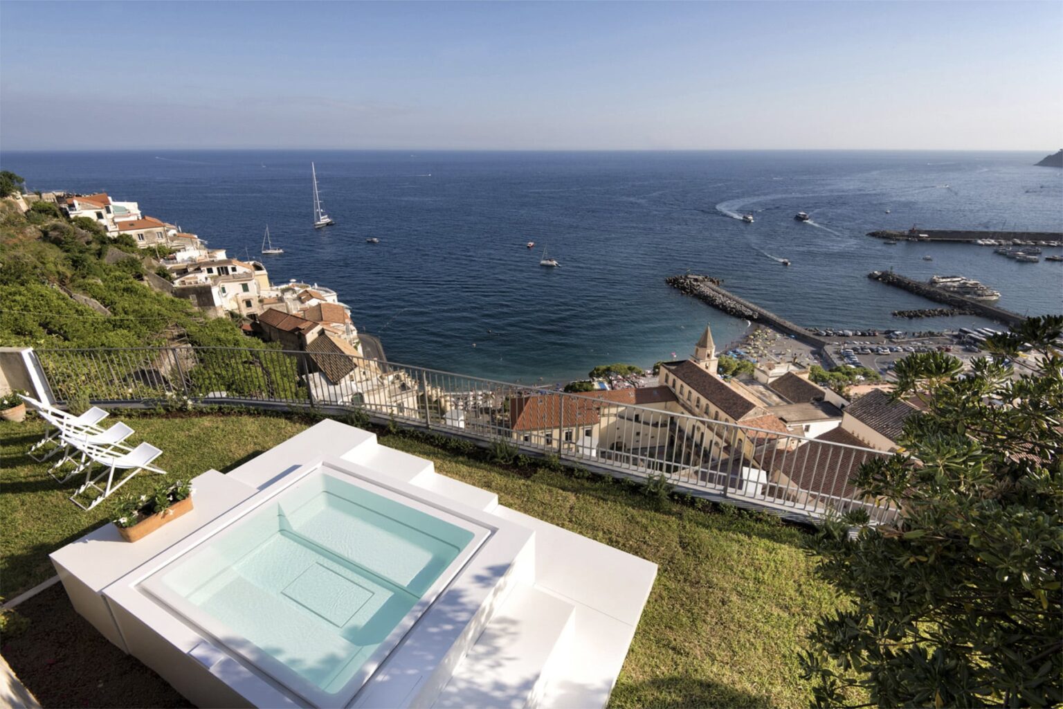 Luxury villa with a private jacuzzi overlooking a scenic coastal view, featuring a marina and sailboats in the distance, surrounded by lush greenery and hillside homes. Perfect for vacation rentals and seaside getaways. | Only on SopranoVillas