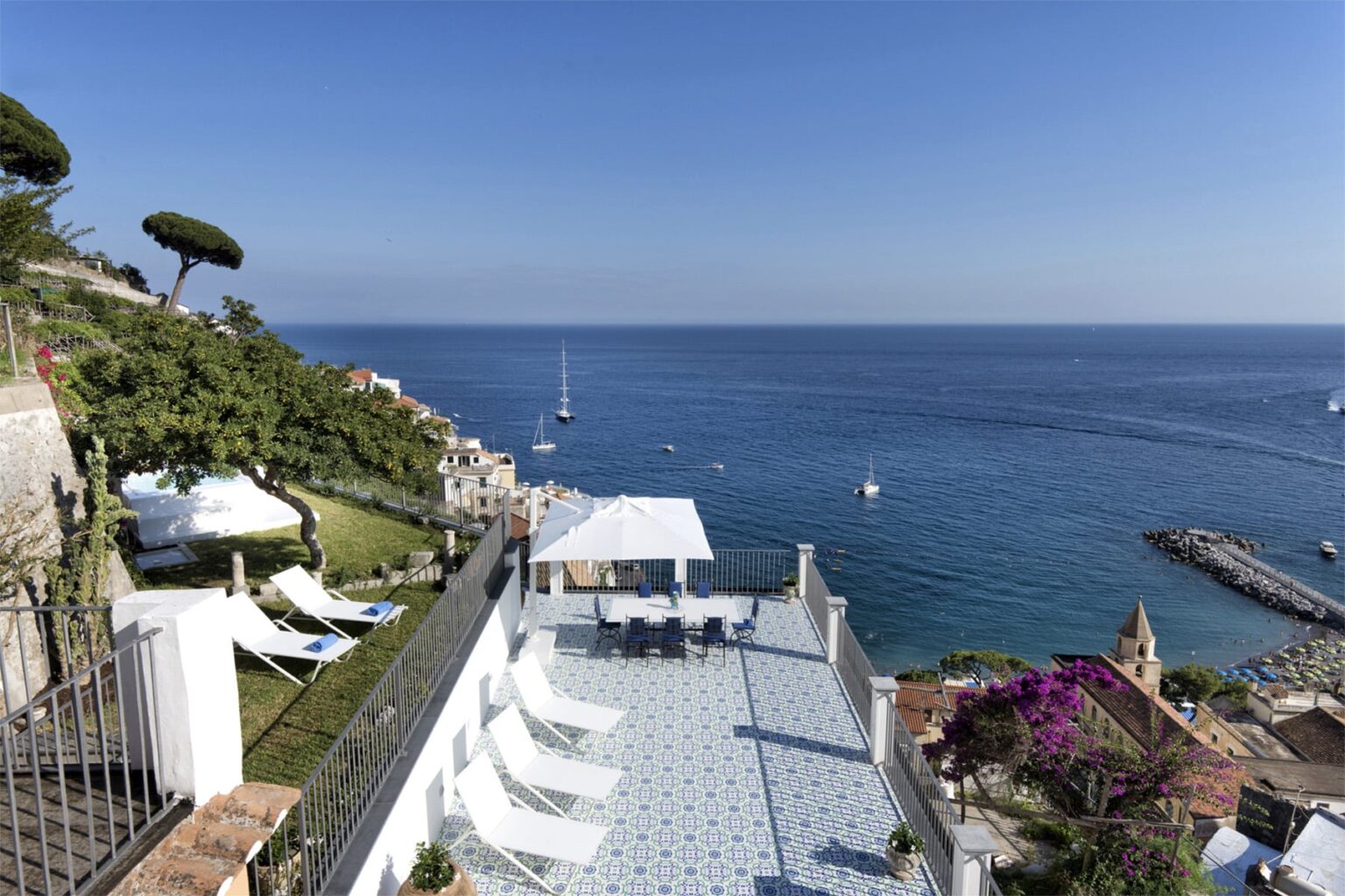 Stunning coastal view from a terrace overlooking the calm blue sea, with sailboats in the distance and lush greenery surrounding the area. Perfect for luxury vacation rentals or seaside retreats. | Only on SopranoVillas