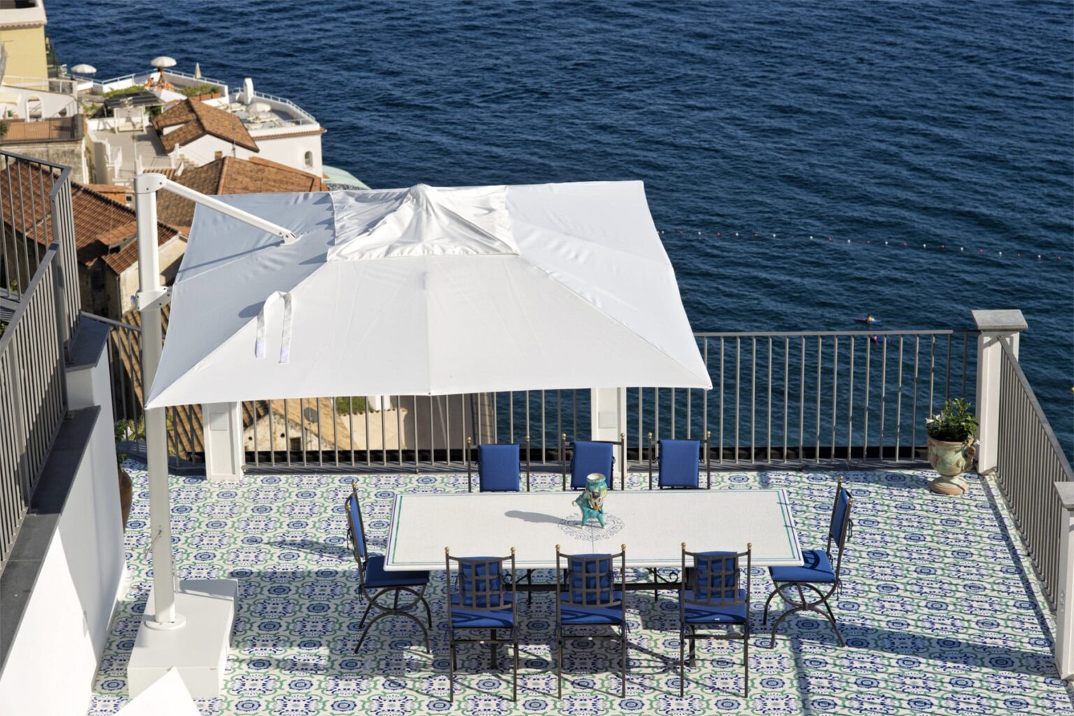 Outdoor dining area with a white umbrella and blue chairs overlooking the sea, featuring a patterned tiled floor and scenic coastal views. | Only on SopranoVillas