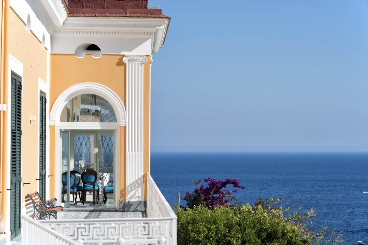 Elegant seaside villa with a yellow exterior, featuring a spacious balcony overlooking the tranquil blue ocean. The balcony is furnished with a table and chairs, surrounded by lush greenery and vibrant flowers, creating a picturesque coastal retreat. | Only on SopranoVillas