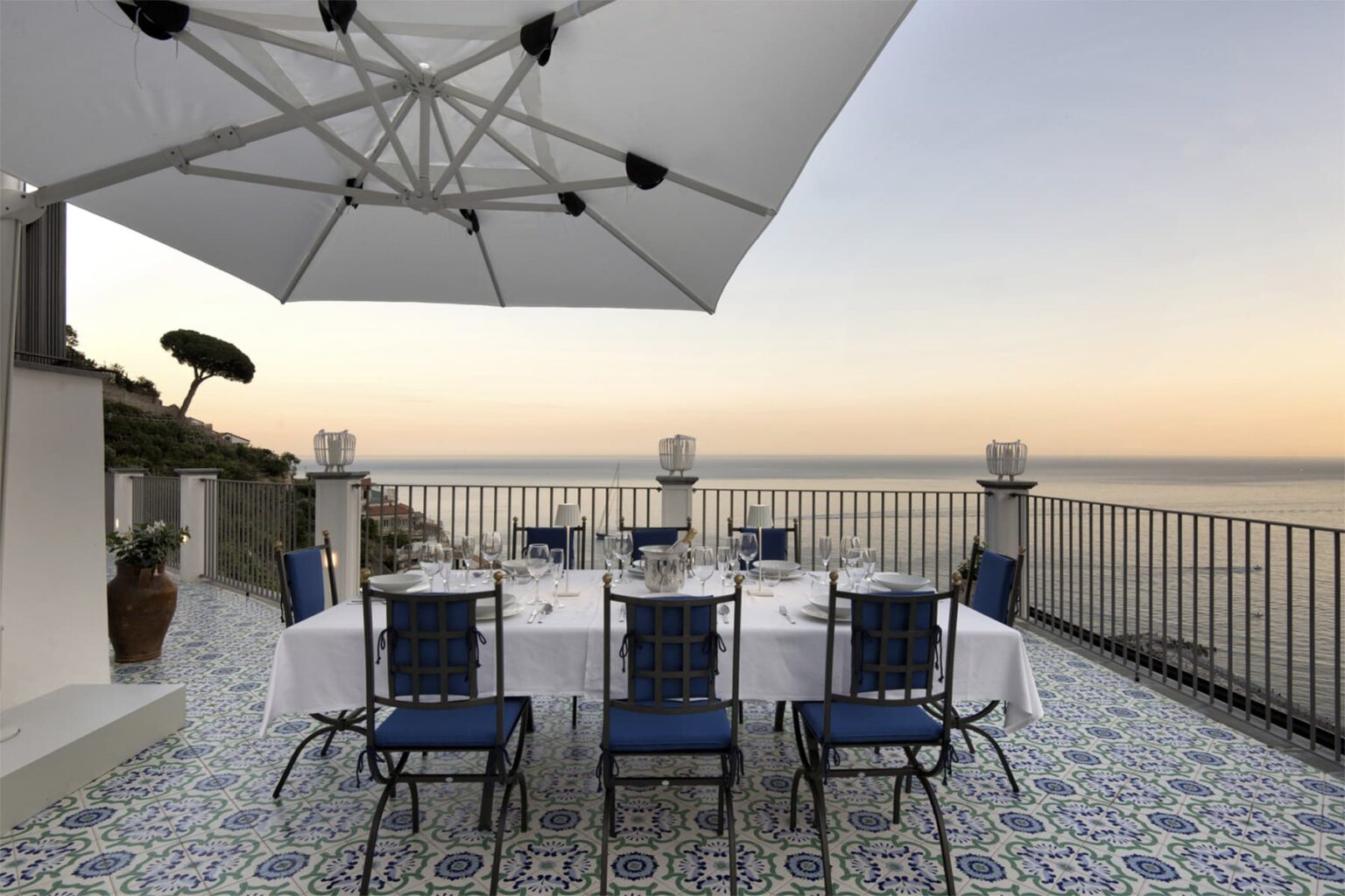 Elegant outdoor dining area with a round table set for eight, featuring blue chairs, white tablecloth, and an umbrella, overlooking a scenic ocean view during sunset. | Only on SopranoVillas