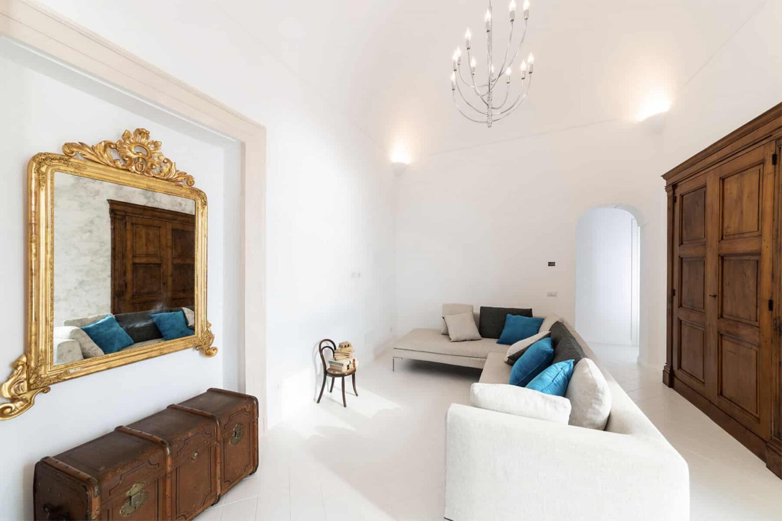 Bright and airy living room featuring a white color palette, elegant chandelier, vintage wooden wardrobe, and a stylish mirror with a gold frame. Comfortable seating adorned with turquoise cushions enhances the sophisticated decor. | Only on SopranoVillas