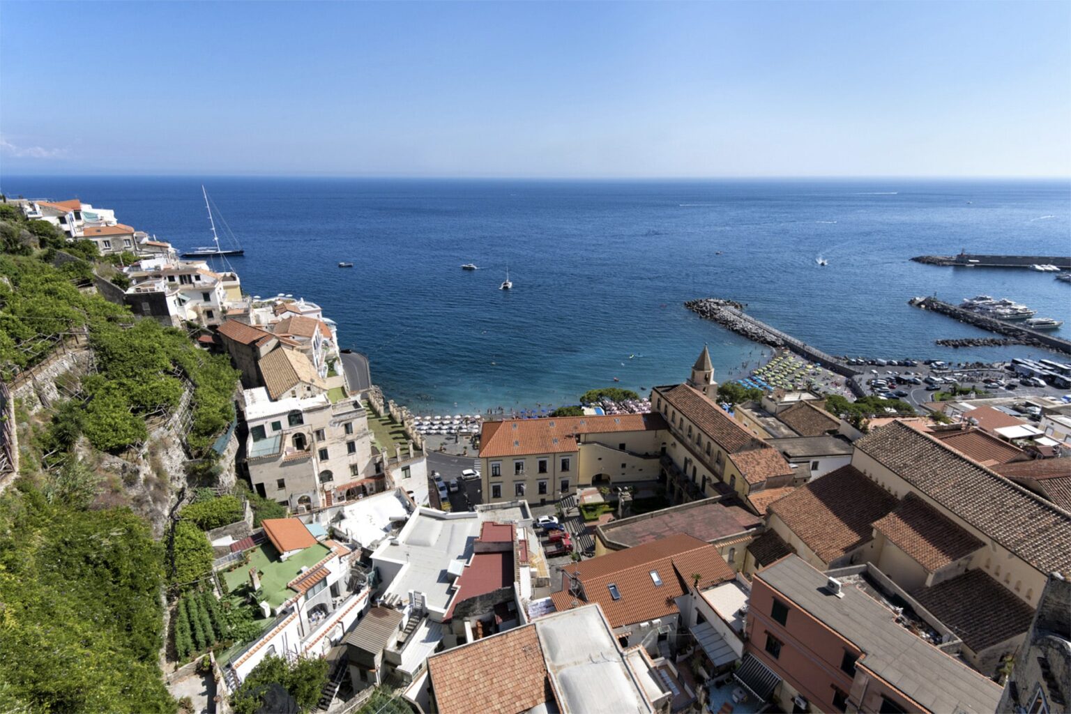 A panoramic view of a coastal town featuring colorful buildings on a hillside overlooking a serene blue sea, with boats in the water and a marina in the distance. The scene captures the charm of Mediterranean architecture and the beauty of the coastline. | Only on SopranoVillas