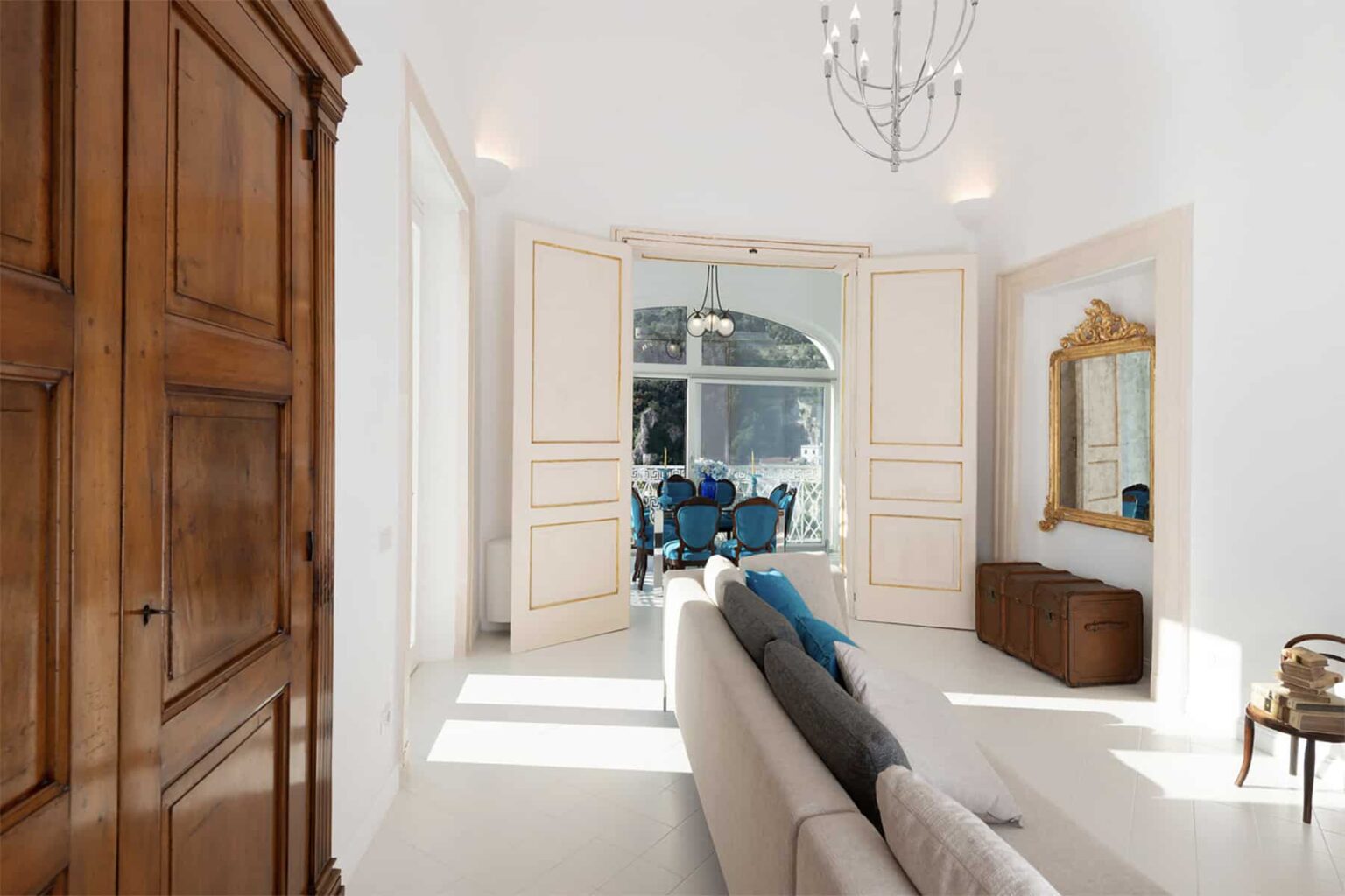 Bright and airy living room featuring a modern sofa, wooden wardrobe, and decorative mirror, with large windows allowing natural light and a view of stylish blue chairs in the background. | Only on SopranoVillas